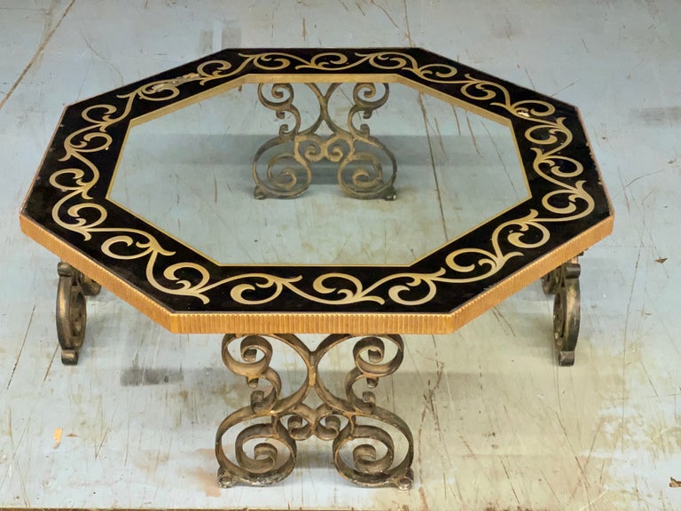 Late 1950's early 1960's Hollywood Regency cocktail table of octagonal form with clear inset glass with gold on black reverse paint eglomise decorative border set within a ribbed and gilt metal frame which is supported by four double-scrolled cast