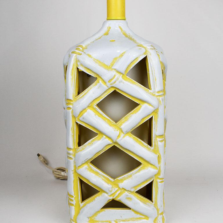 A ceramic faux bamboo lamp in white and yellow. This piece is square with rounded corners. Faux bamboo shoots criss cross at the sides showing the pendant globe inside. The top is white, with yellow bamboo shoots at each corner. A wooden finial in