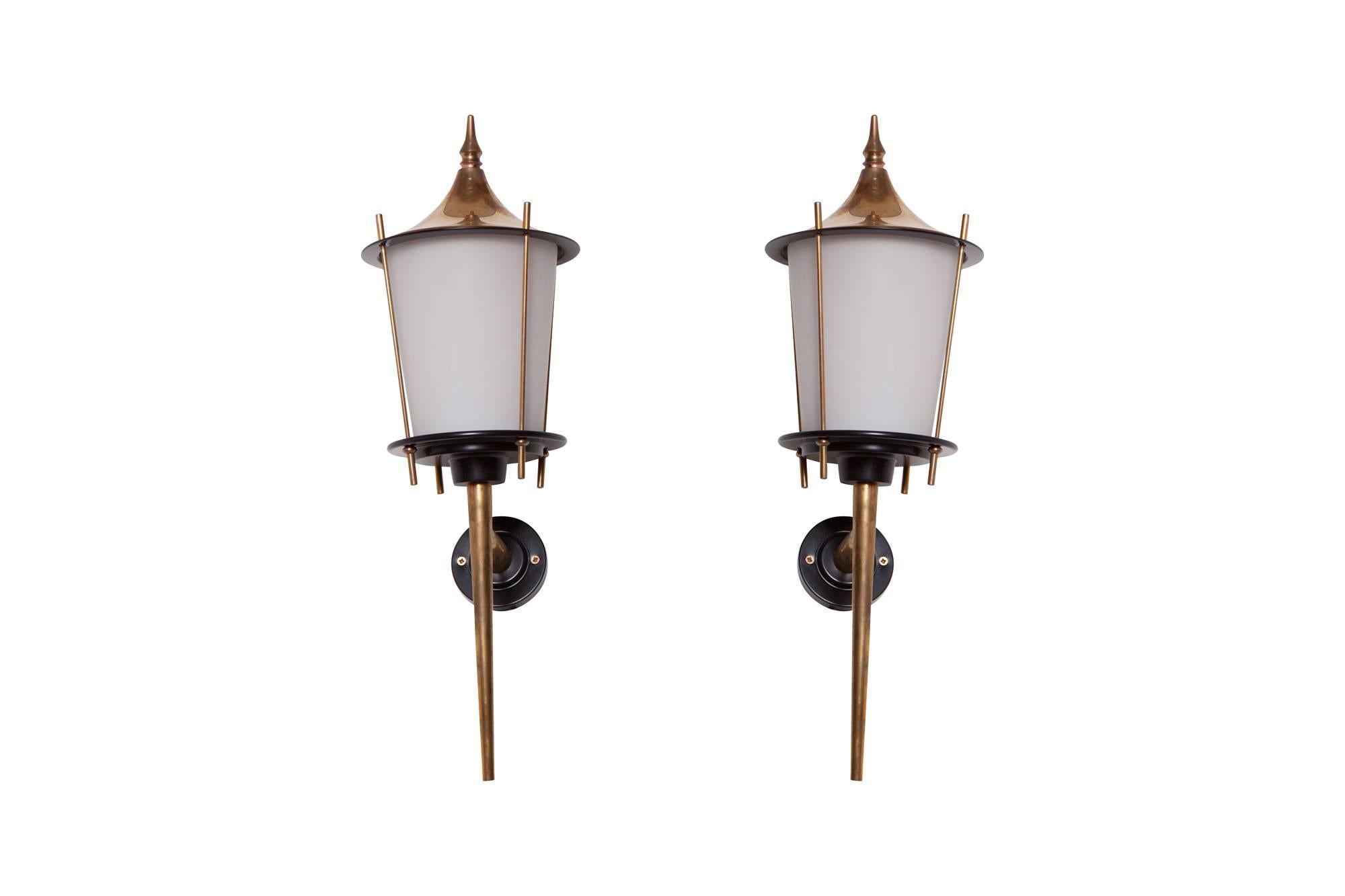 Mid-century modern brass Lantern shaped wall lights by Maison Arlus in brass and black lacquered metal, France, 1970s.
The opaline glass shades provide the light with an elegant and soft light partition. The lights are finished with brash hats to