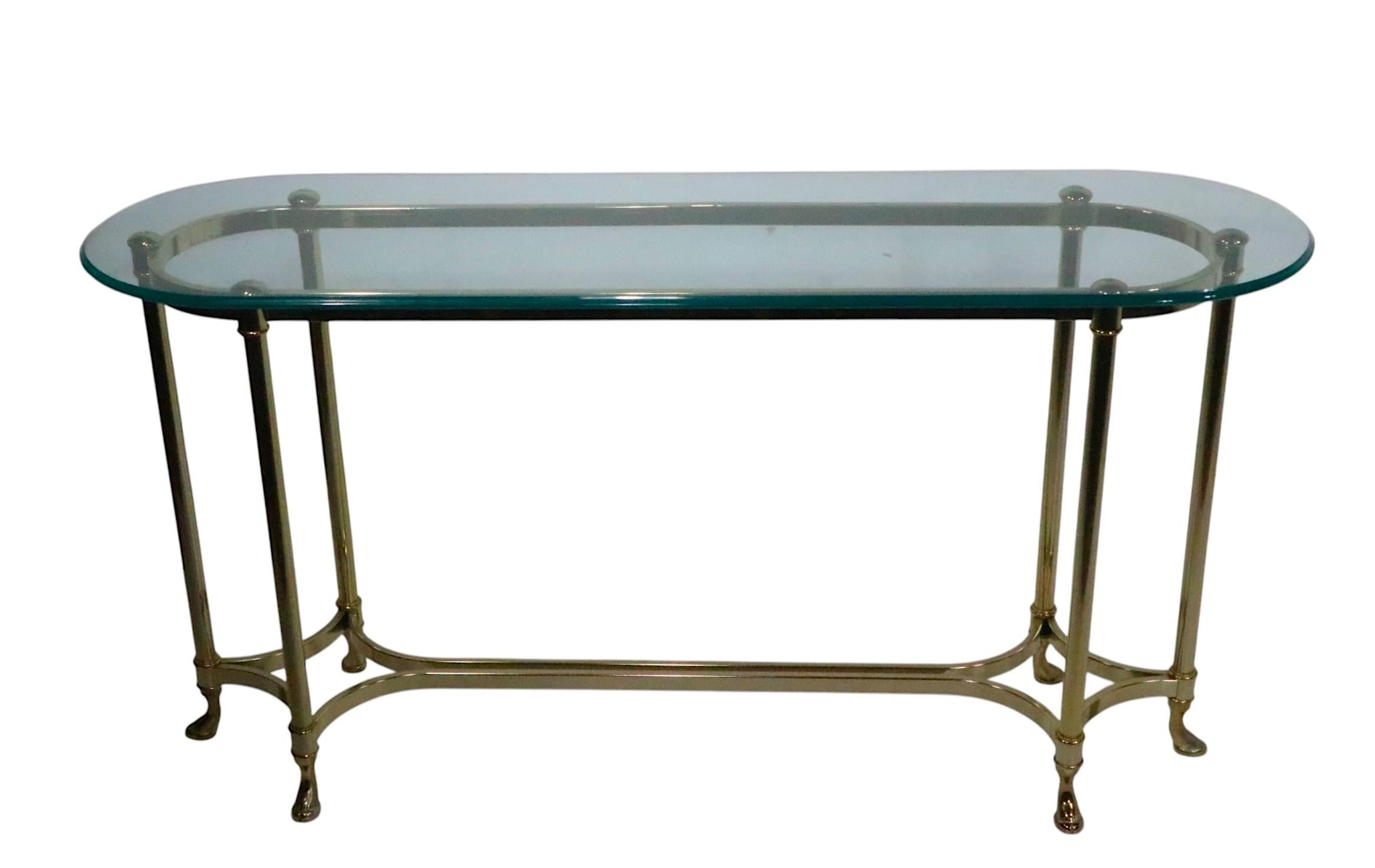 Chic Hollywood Regency, Palm Beach Modern style glass top console table. The oval glass top rests on a stylized brass plate metal base. This example is in good, original, clean and ready to use condition, showing only light cosmetic wear, normal and