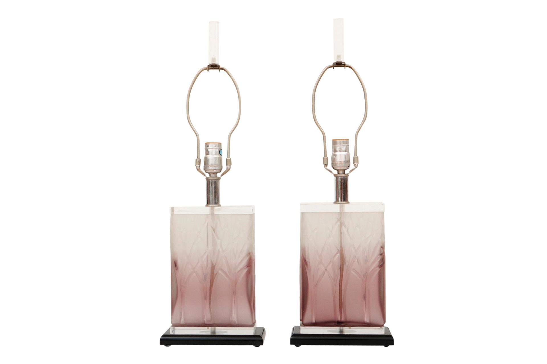 A pair of 1980’s Hollywood Regency lucite table lamps. Blocks of translucent lucite are rich purple fading to white and pressed with trees in the front and back. Short chrome columns support sockets, harps and cylindrical clear lucite finials.