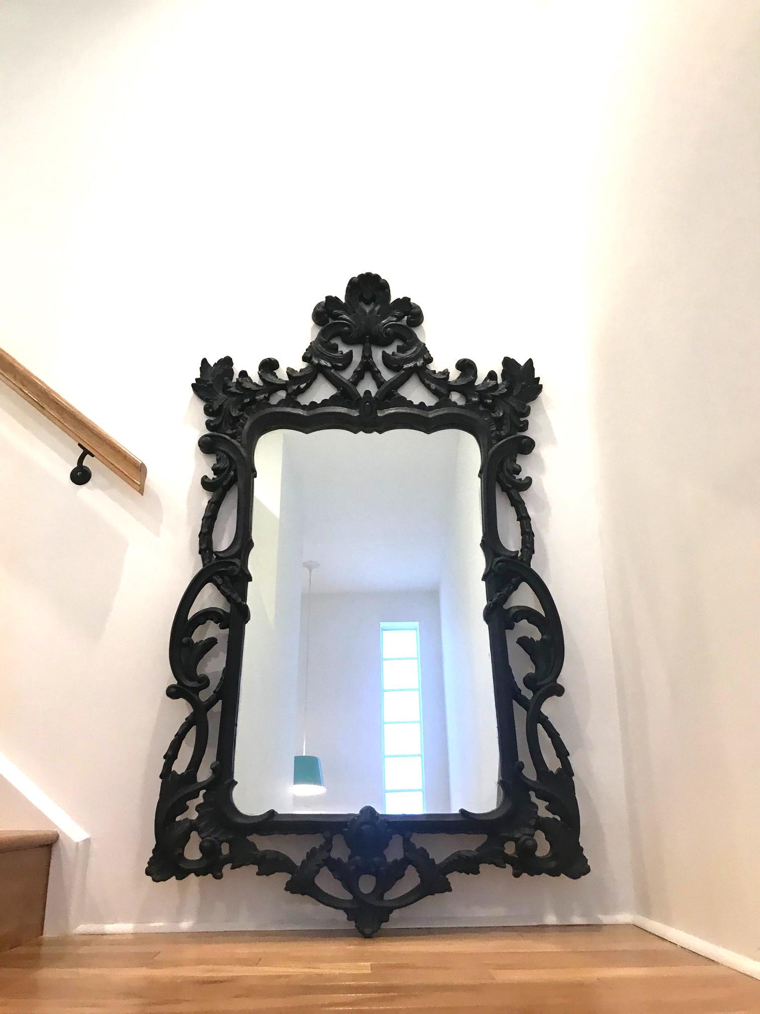 Large Hollywood Regency mirror with traditional Rococo style carved wood frame. Mirror has shield design featuring a series of hand carved scrolls with foliage details. The vintage black finish has a slightly distressed quality which gives the