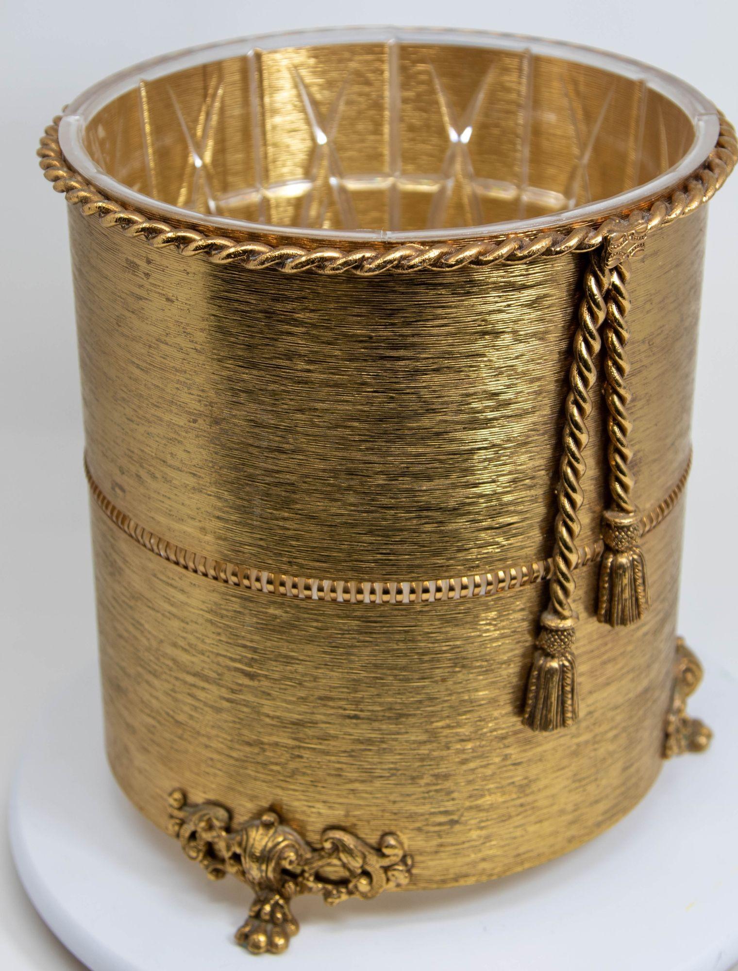Vintage Mid Century Hollywood Regency Glam rope and tassel wastebasket with claw feet and original clear plastic liner.
This Hollywood Regency is crafted from metal brushed brass with a gilt gold ormolu finish with cast brass details featuring an