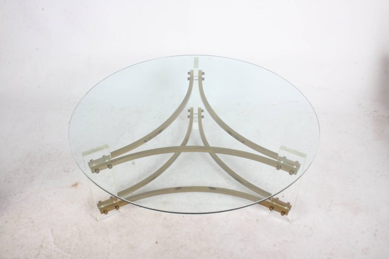 Circa 1970s Hollywood Regency style round coffee table with Lucite legs and brass support bars. 
Brass has patina, can be polished for addition cost. Lucite and glass have light scuffs. 