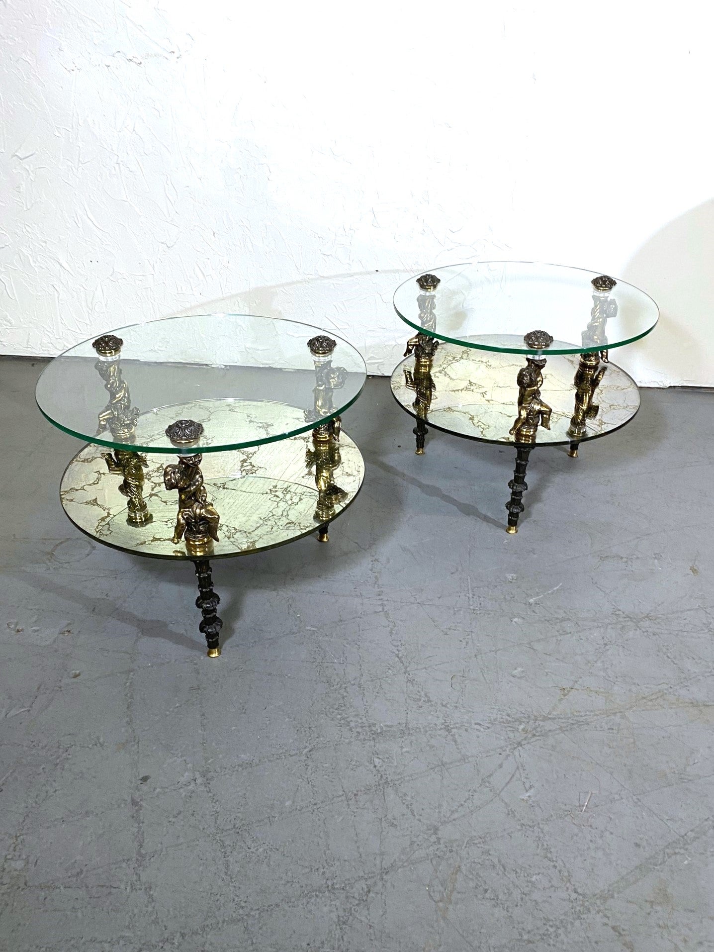 Paire de tables d'appoint à deux niveaux de forme ronde de style Hollywood Regency. La table a trois pieds avec des chérubins en laiton sur chacun d'eux.