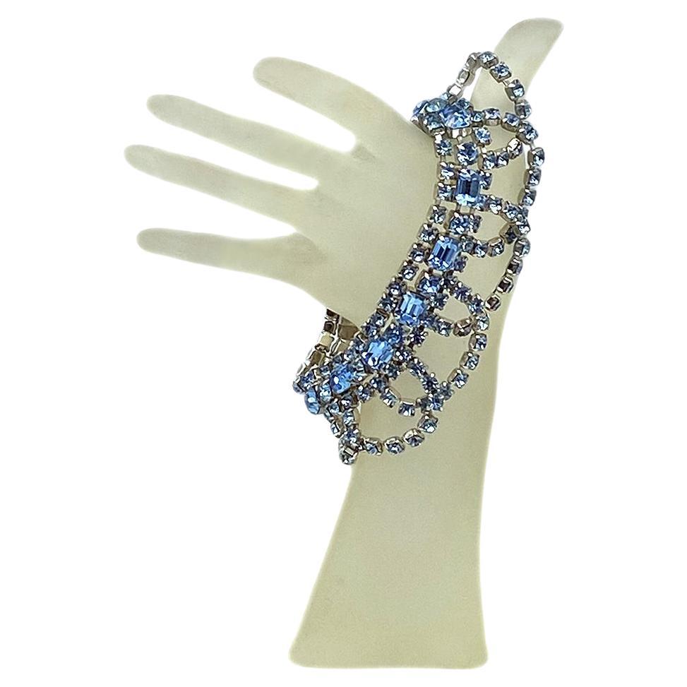 This is a Hollywood Regency style French blue garnished bracelet. It has prong set round and emerald cut rhinestones on rhodium plated metal. This high end vintage bracelet is garnished with overlapping swags and comes with a security chain. It will