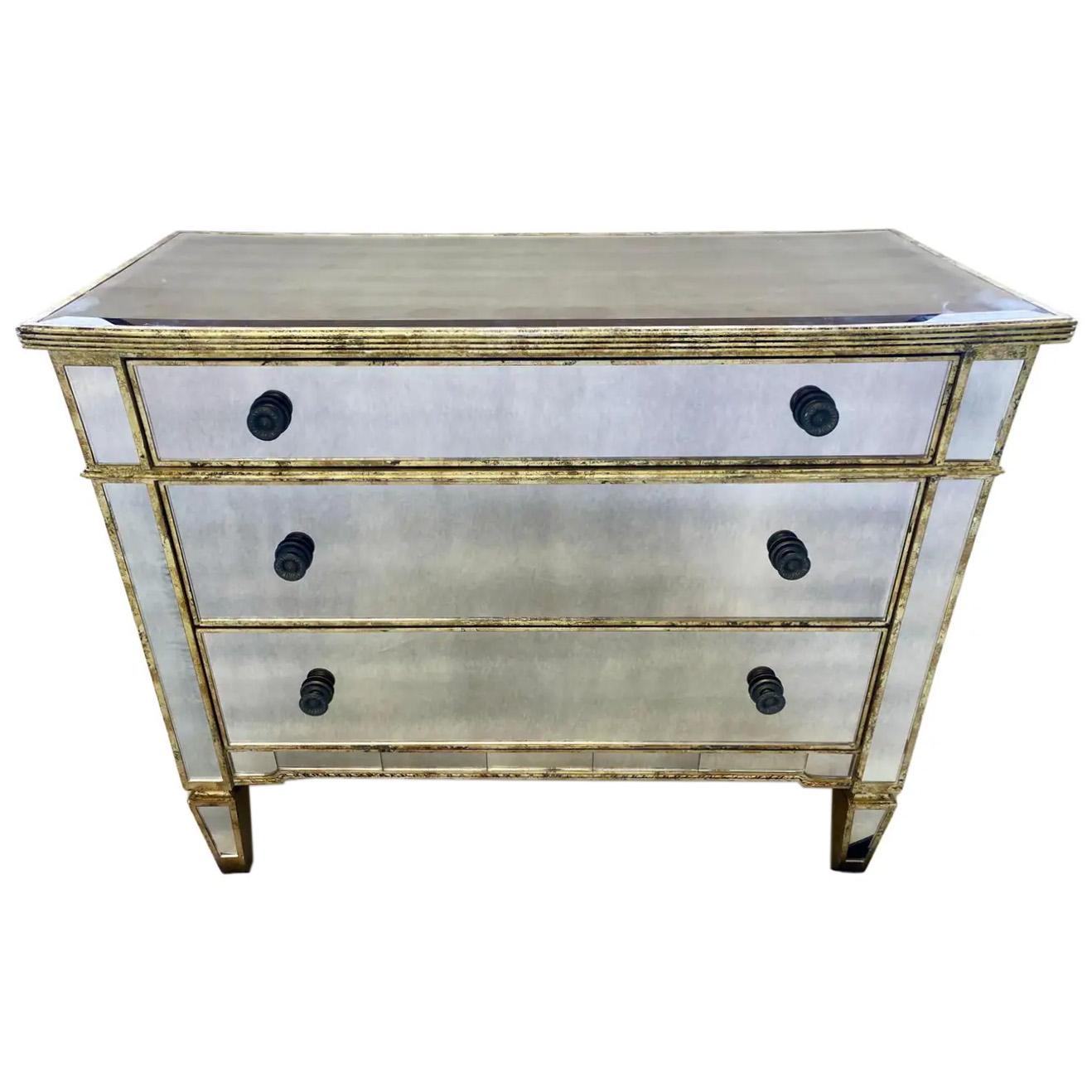 A quality Hollywood Regency style three drawer commode, dresser or chest. The commode features distressed mirrored glass and is embellished with gold Champagne trim or frame also in an antiqued finish with leaf pattern. 
The beautiful commode or