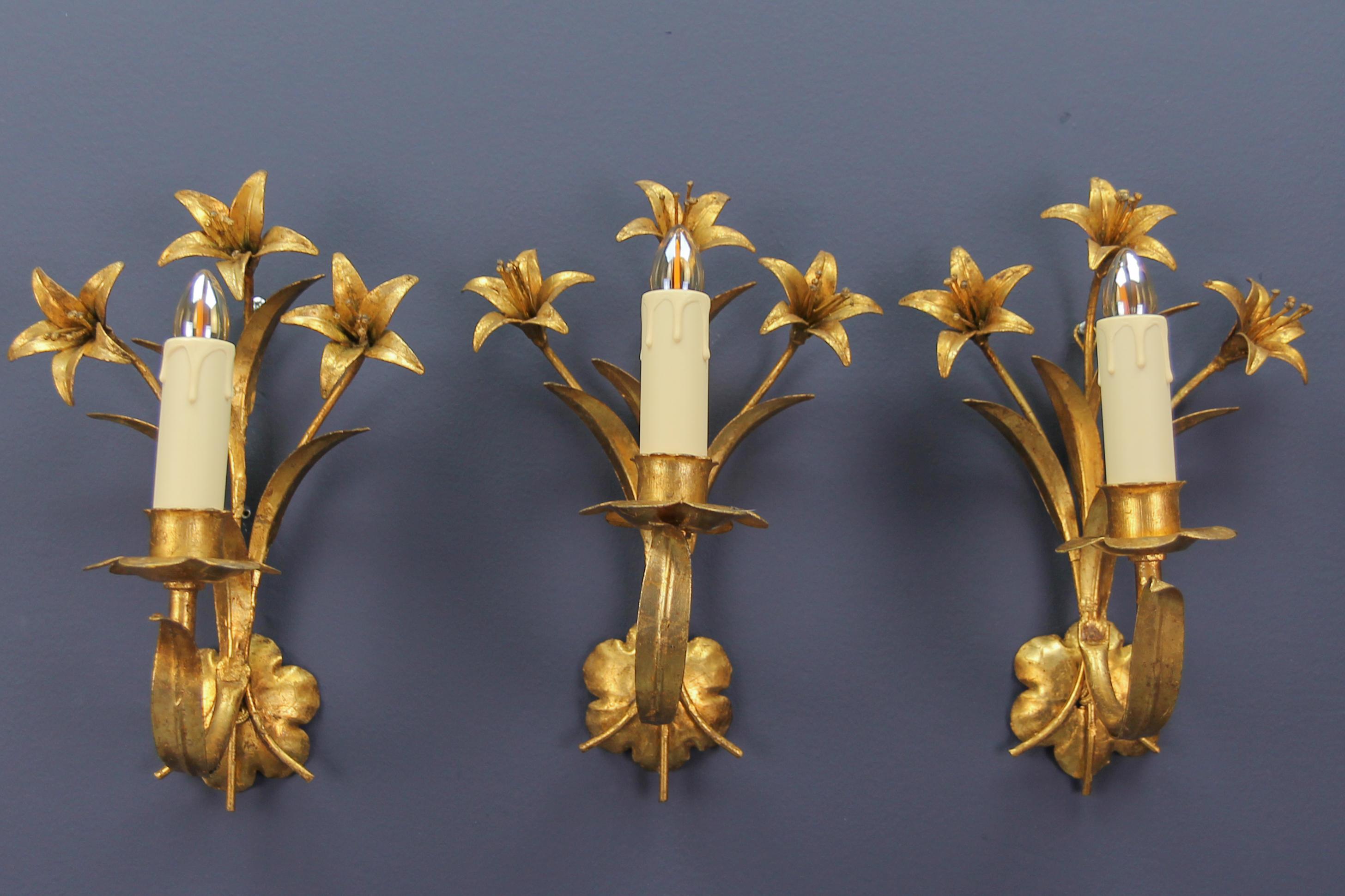 Hollywood Regency style French gilt metal lily flower wall sconces, set of three.
A beautiful set of three gilt metal wall lights in the shape of lilies. Each sconce has one arm with a socket for an E14 size light bulb and three flowers and leaves