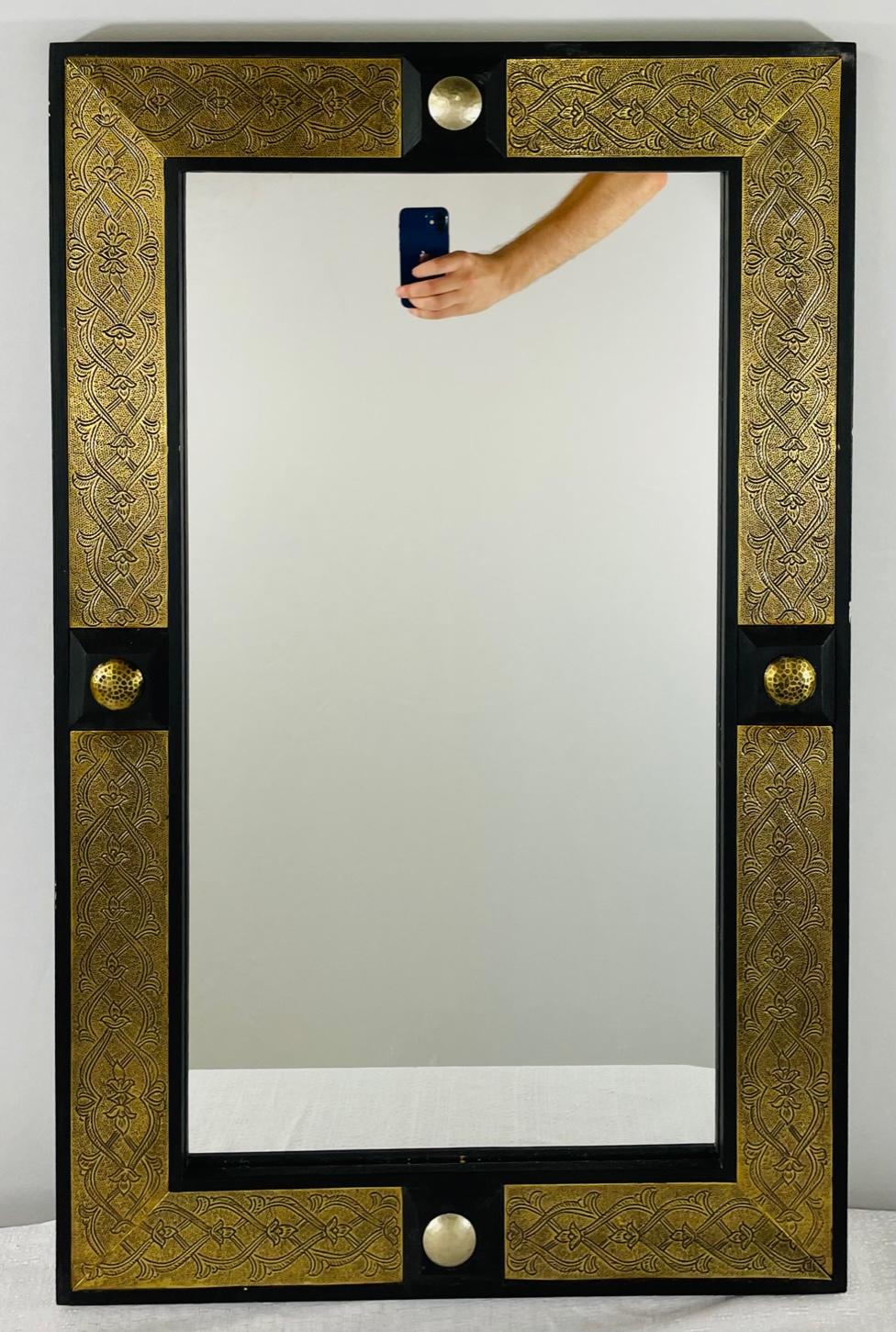 Hollywood Regency style Moroccan mirror ebonized on brass

The mirror has a rectangular frame of ebonized wood flanking a brass inset hand designed in filigree style with ebony backed brass button decorations. The mirrors can be hung vertically or