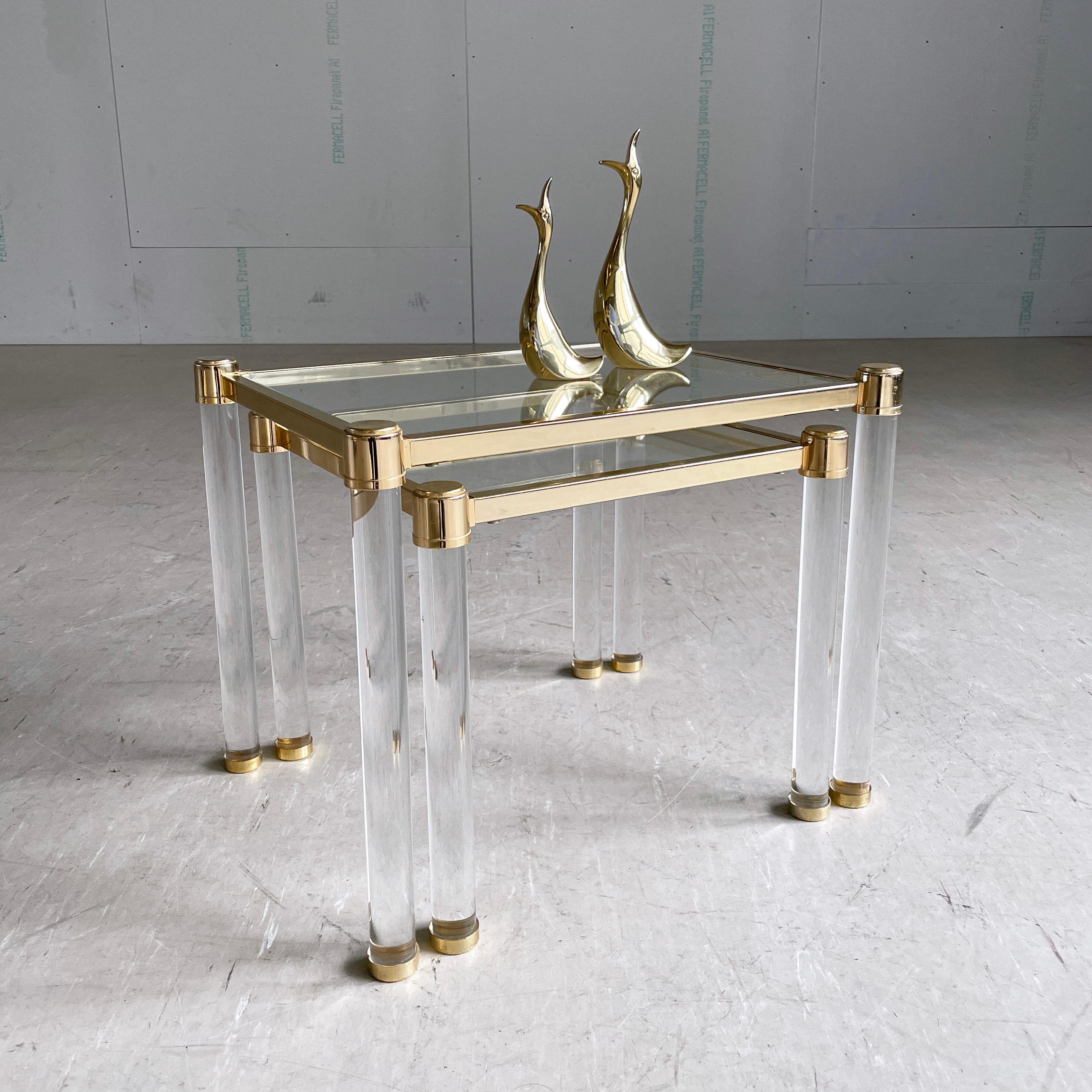 Pair of Hollywood Regency style nesting tables (set includes 2 tables). Clear glass tabletop with gold metal frame and leg bases. Transparent acrylic Legs.  
Measurements: 
Small Table - Height 36cm Width 40cm Depth 40cm
Large Table - Height 41cm