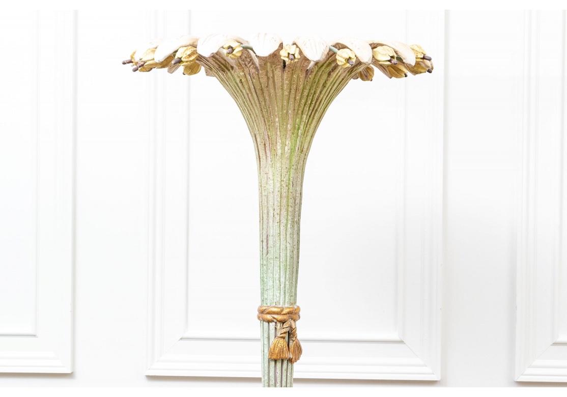 A tall carved ribbed palm tree in antiqued pale green paint. The top pale leaves alternating with metal stemmed off white buds. The trunk with two carved gilt ribbons with tassels. The flared shaped bottom raised on a tiered painted and gilt base.