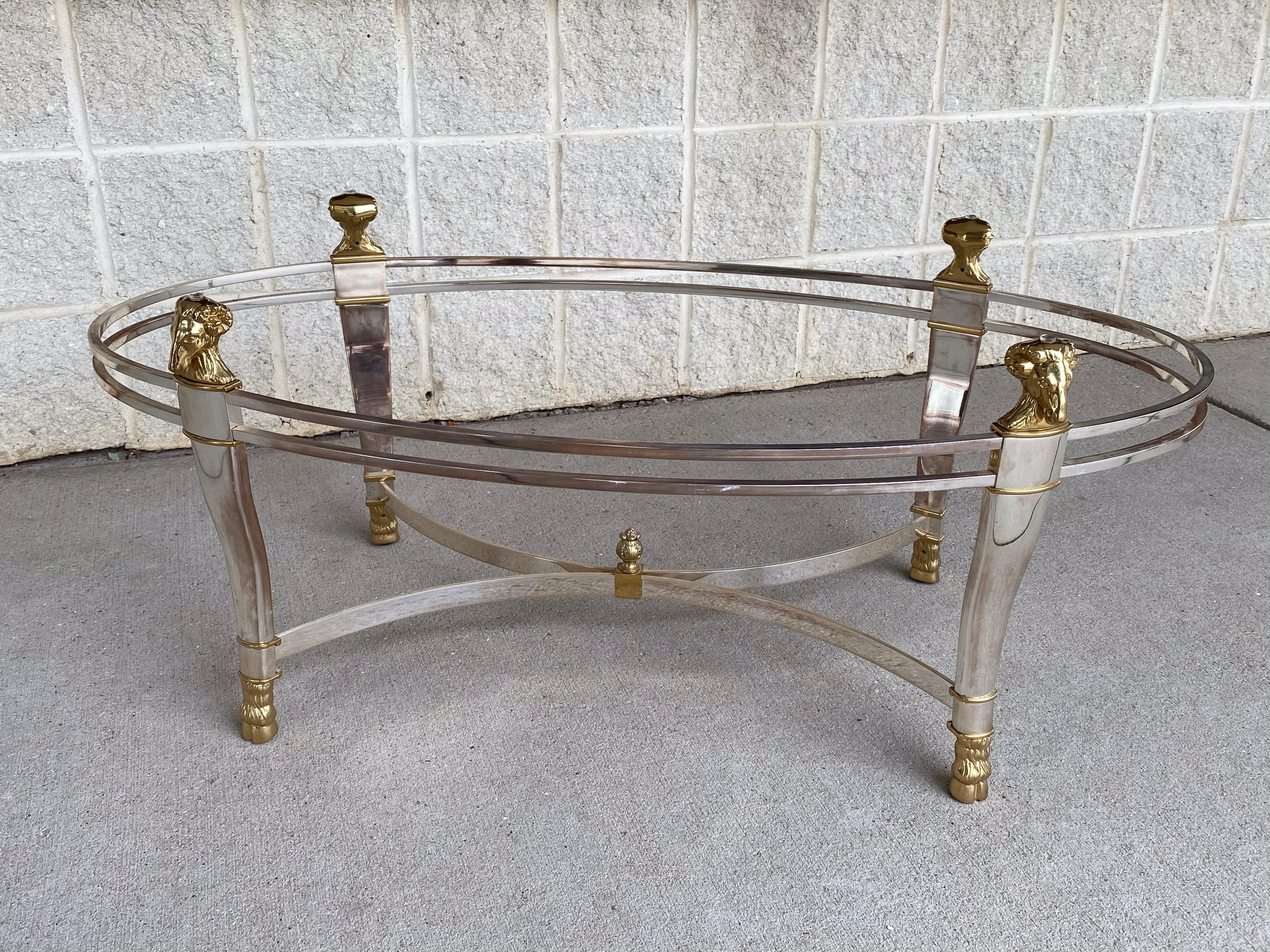 Hollywood Regency style oval or elliptical shaped cocktail or coffee table crafted from solid brass in silver plated finish with polished brass accents. ram's head and hoof details with decorative pom de pin final at stretcher. Oval floating glass