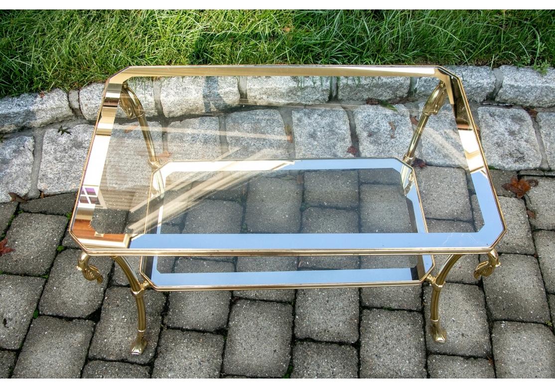 A smaller scale tiered cocktail table with strong proportion and form. Rectangular with canted corners and a glass top and lower shelf with mirrored bands. A fine brass frame with curved legs with swan heads and web feet.
Condition: Acceptable age