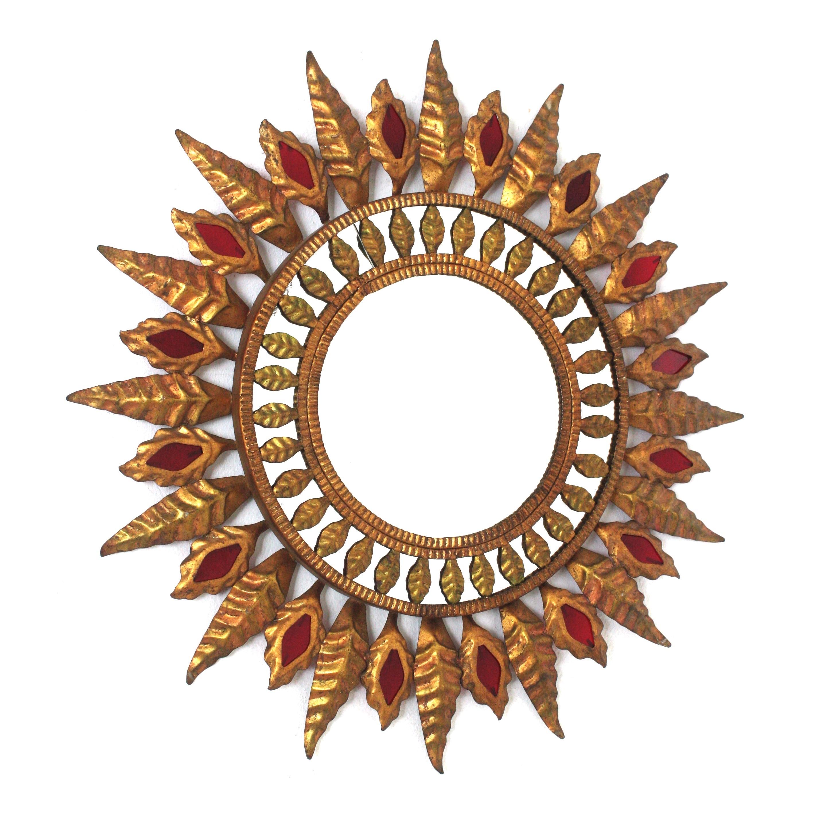 Sunburst Mirror in Gilt Iron and Red Glass, Spain, 1950s.
Eye-catching sunburst mirror featuring a mirrored surface surrounding the central glass, a frame with alternating large and small leaves and red glass accents. 
Nice aged patina and original