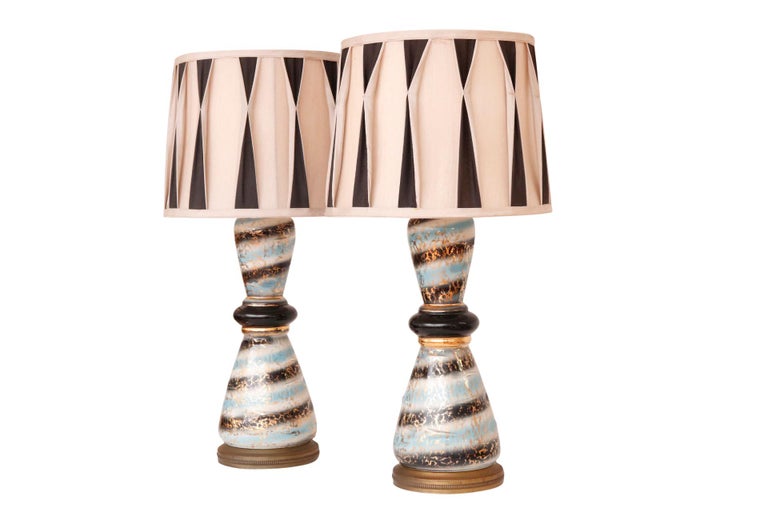 A pair of Hollywood Regency style ceramic table lamps. Custom shades are pleated in cream and black. Hourglass vases are decorated in a blue, white and black swirl glaze with a gilt overlay, finished with round brass bases. Each lamp measures 19.25