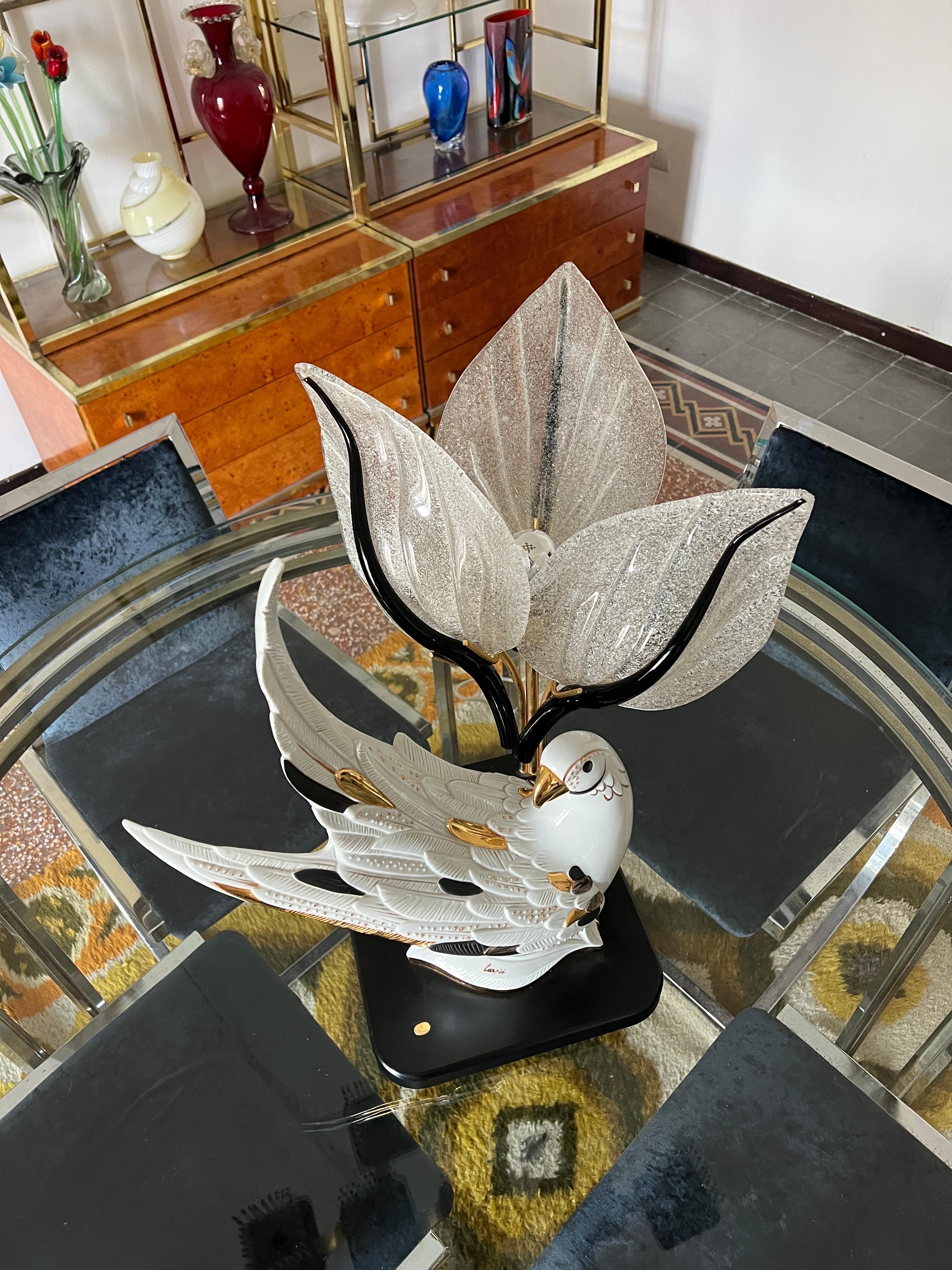 Behold our exquisite 1980s ceramic bird table light. This eye-catching piece features a stunning white ceramic bird adorned with intricate gold and black detailing. Three large Murano glass leaves surround the light socket, crafted using the