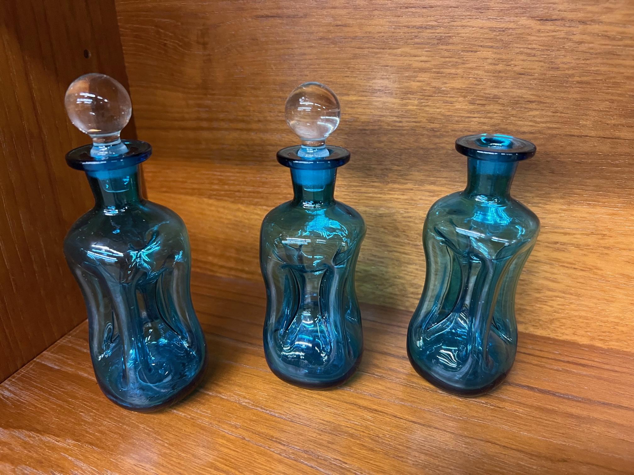 Set of 3 blue glass decanters by Holmegaard. Called Kluk Kluk decanters because of the sound they make when pouring into a cocktail glass. Two with stoppers, one without stopper.
Handblown glass with pinched sides, clear glass contrasting crown
