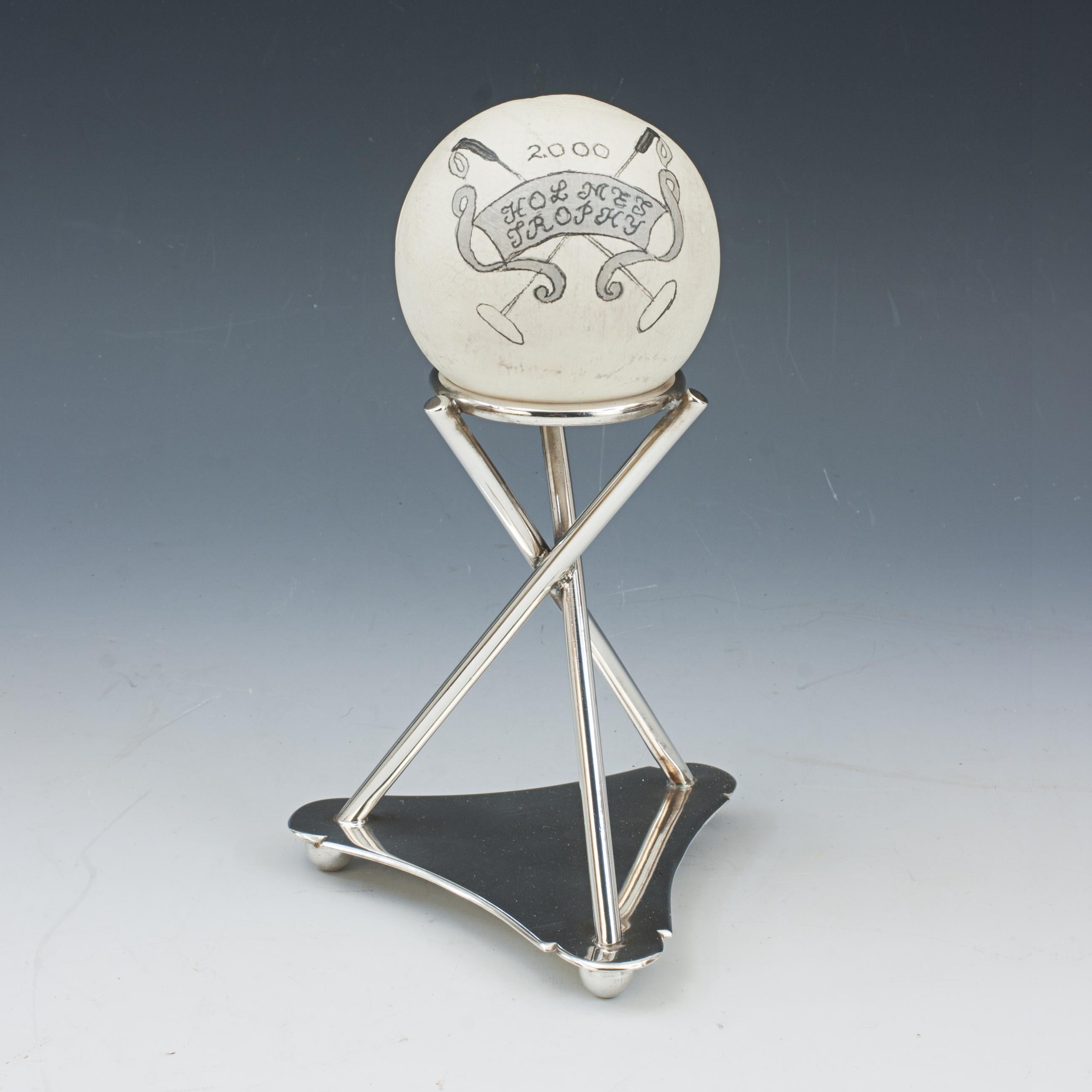 Vintage Polo Trophy.
A white wooden polo ball on a plated tripod stand. The polo ball painted with two crossed polo mallets, a banner reading 'HOLMES TROPHY, 2000'. The base with a flat shaped triangular plate with three ball feet, three raised bars