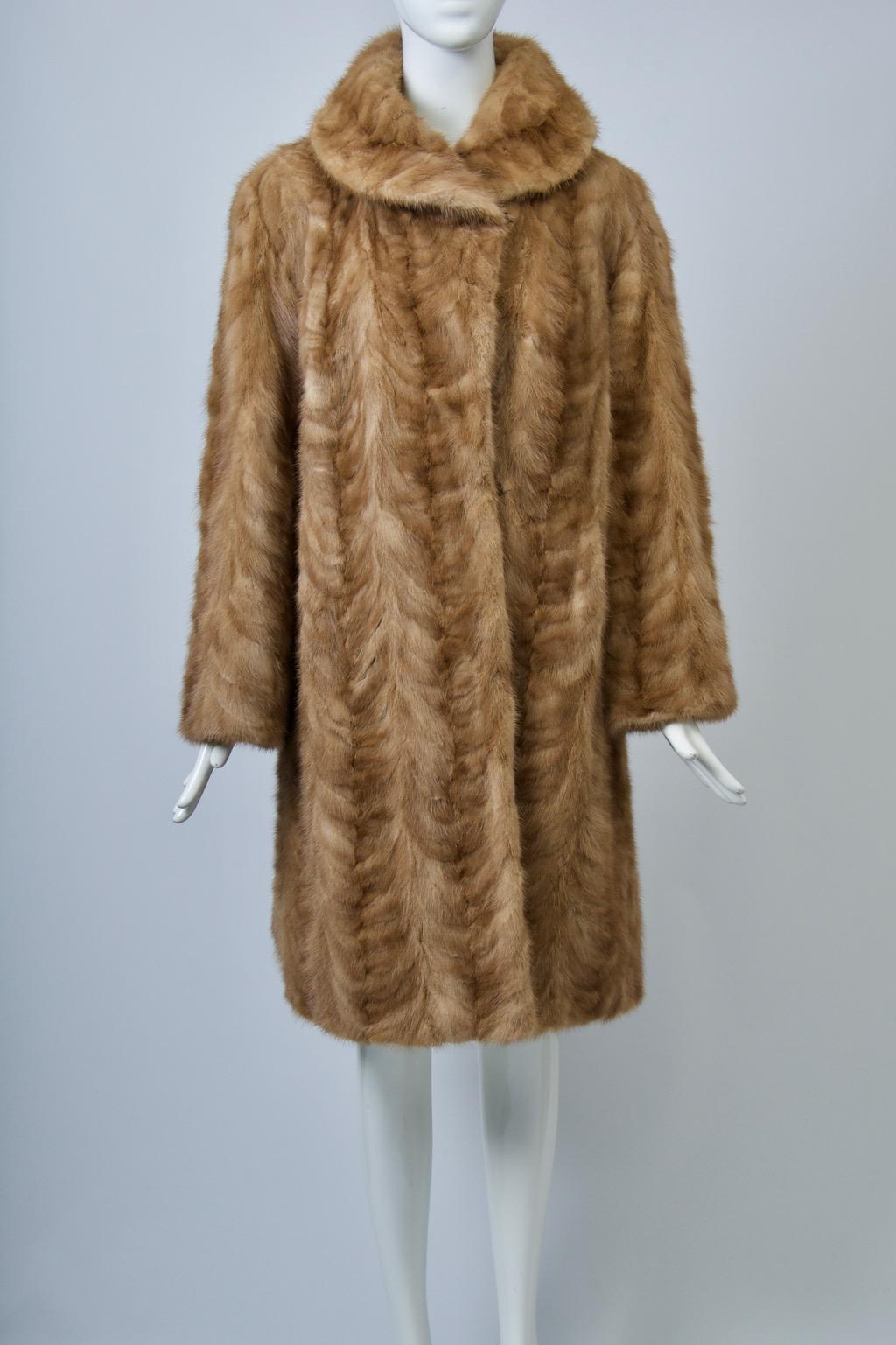 Le manteau en vison couleur miel, avec un motif à chevrons, présente une silhouette droite avec un peu d'ampleur dans le dos, des manches droites rentrées et un col arrondi, ainsi que des poches en biais. L'intérieur est doté d'une doublure brodée