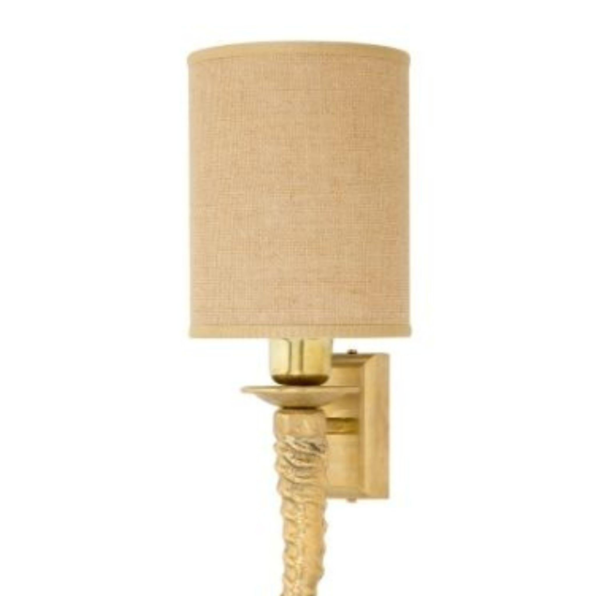 Lámpara de pared con estructura de latón fundido en forma de cuerno y pequeña pantalla cilíndrica de tejido suelto. Este aplique es perfecto para un interiorismo de estilo animalier y selvático, un mueble ecléctico en el que la artesanía se mezcla
