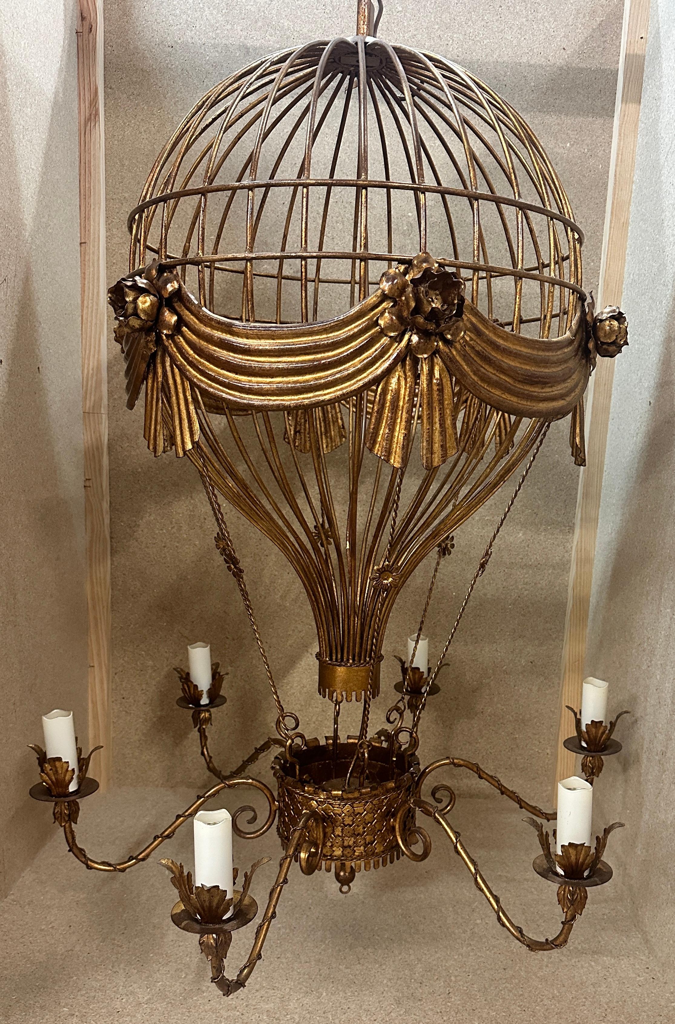 Unusual and whimsical gilt and bronzed tole hot air balloon form chandelier. Balloon adorned with draped swags and roses. Basket below suspended in realistic form from wires as ropes. Six arms/lights projecting from basket.