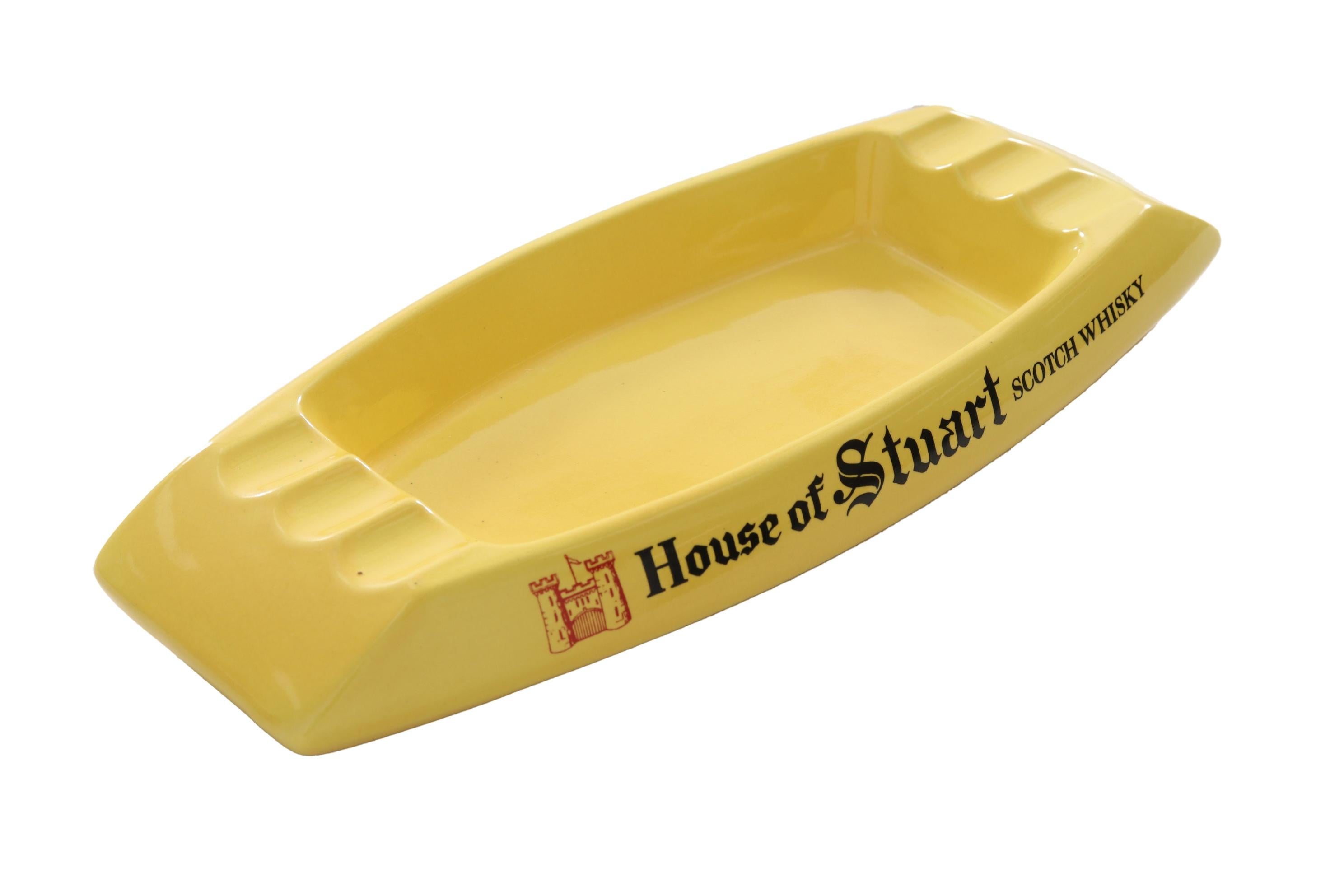 A ceramic House of Stuart Scotch Whisky branded cigar ashtray. Bright yellow and shaped like a boat, the ashtray has three cigar rests at each end and is branded with the House of Stuart name on each side. Marked 
