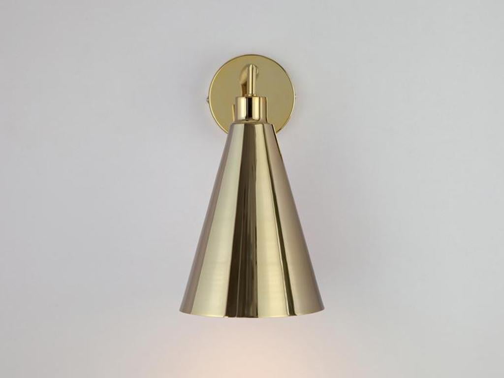 This sleek and practical wall sconce fuses traditional and contemporary. The stem base suspends an oversized cone shade which casts a wide atmospheric glow. Ideal for a living space or bedside light. Rich and opulent, Brass is the houseof staple