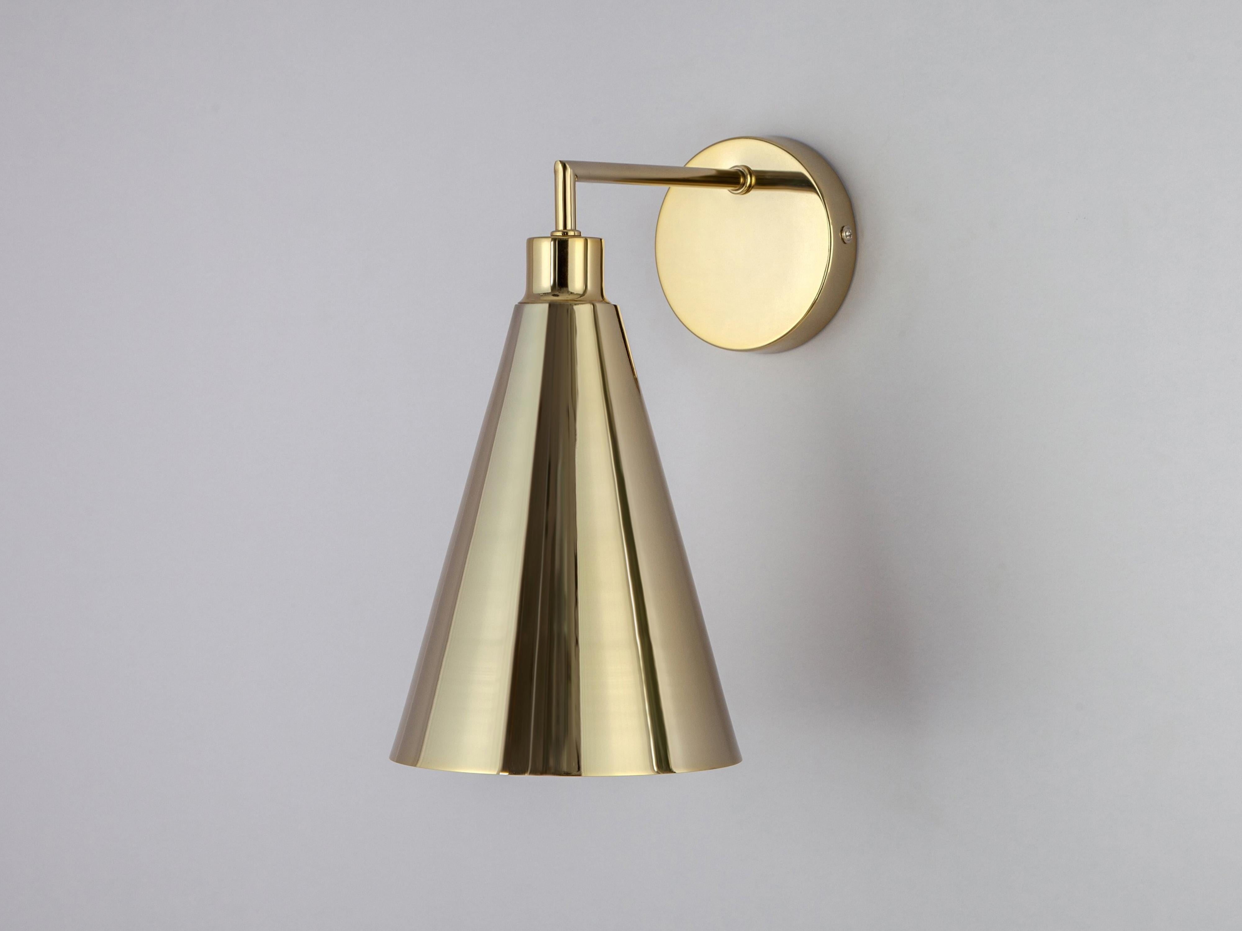 This sleek and practical wall sconce fuses traditional and contemporary. The stem base suspends an oversized cone shade which casts a wide atmospheric glow. Ideal for a living space or bedside light. Brass brings depth and opulence to your interior