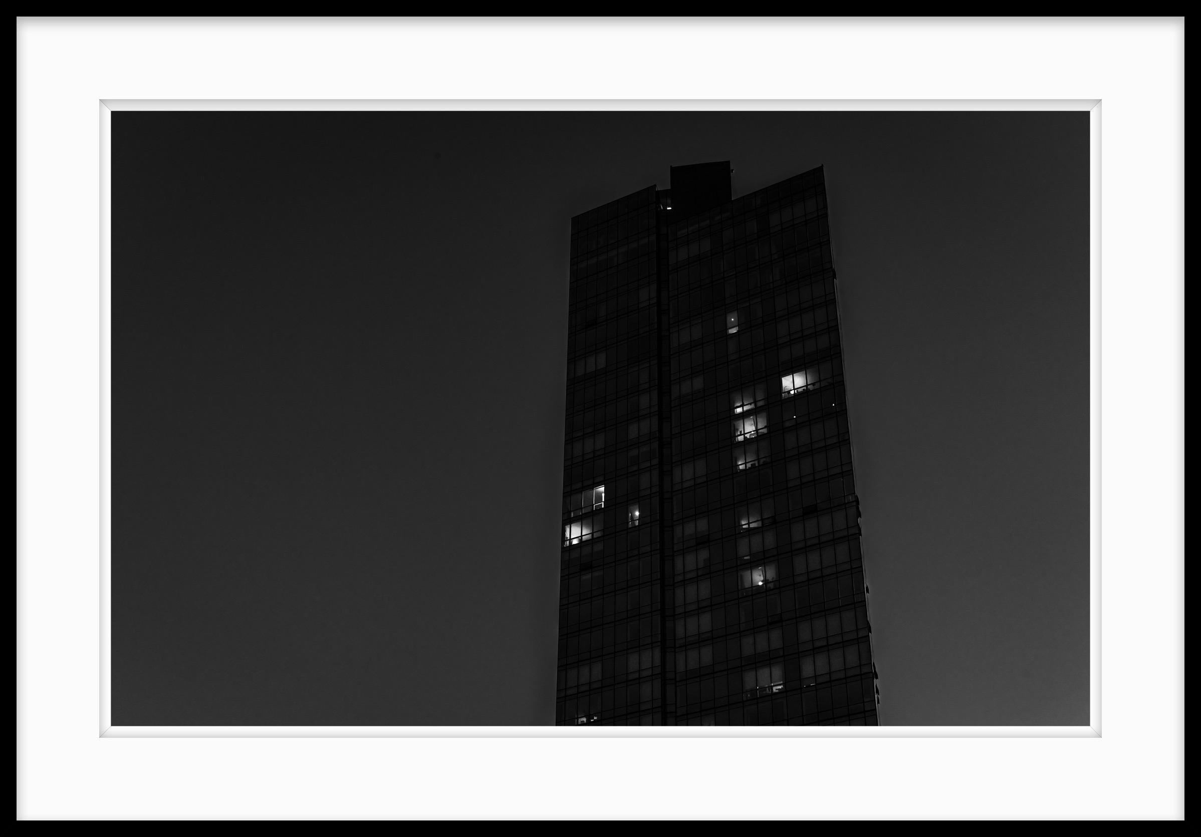 This is a limited edition black and white photograph called Working Late taken in 2021. It captures, at the height of the Covid epidemic, the changing nature of where and how we work. Office buildings are relatively empty as night falls but a few