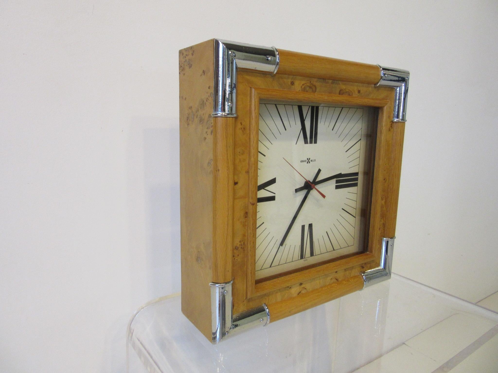 A maple burl wood framed wall clock with cast chrome accents to each corner and a cream face with roman numeral numbers. Battery operated and hanging hole drilled to the backside with paper information certificate from the manufacture Howard Miller