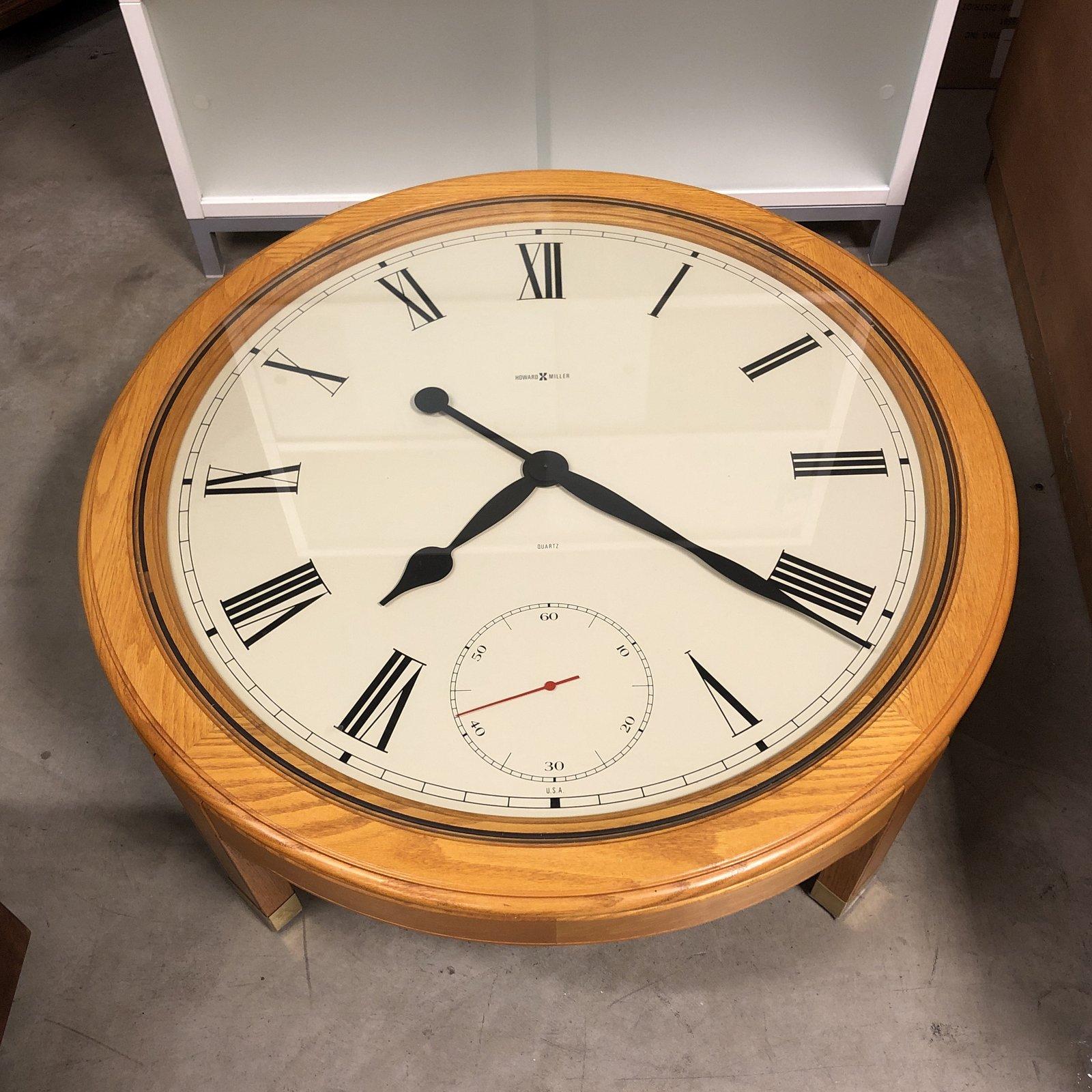 We presents a clock coffee table by Howard Miller Clock Company. A clock face cocktail table, circular with an oak frame. A white face with black Roman numerals and hands, a glass top with brass capped feet. Battery operated.