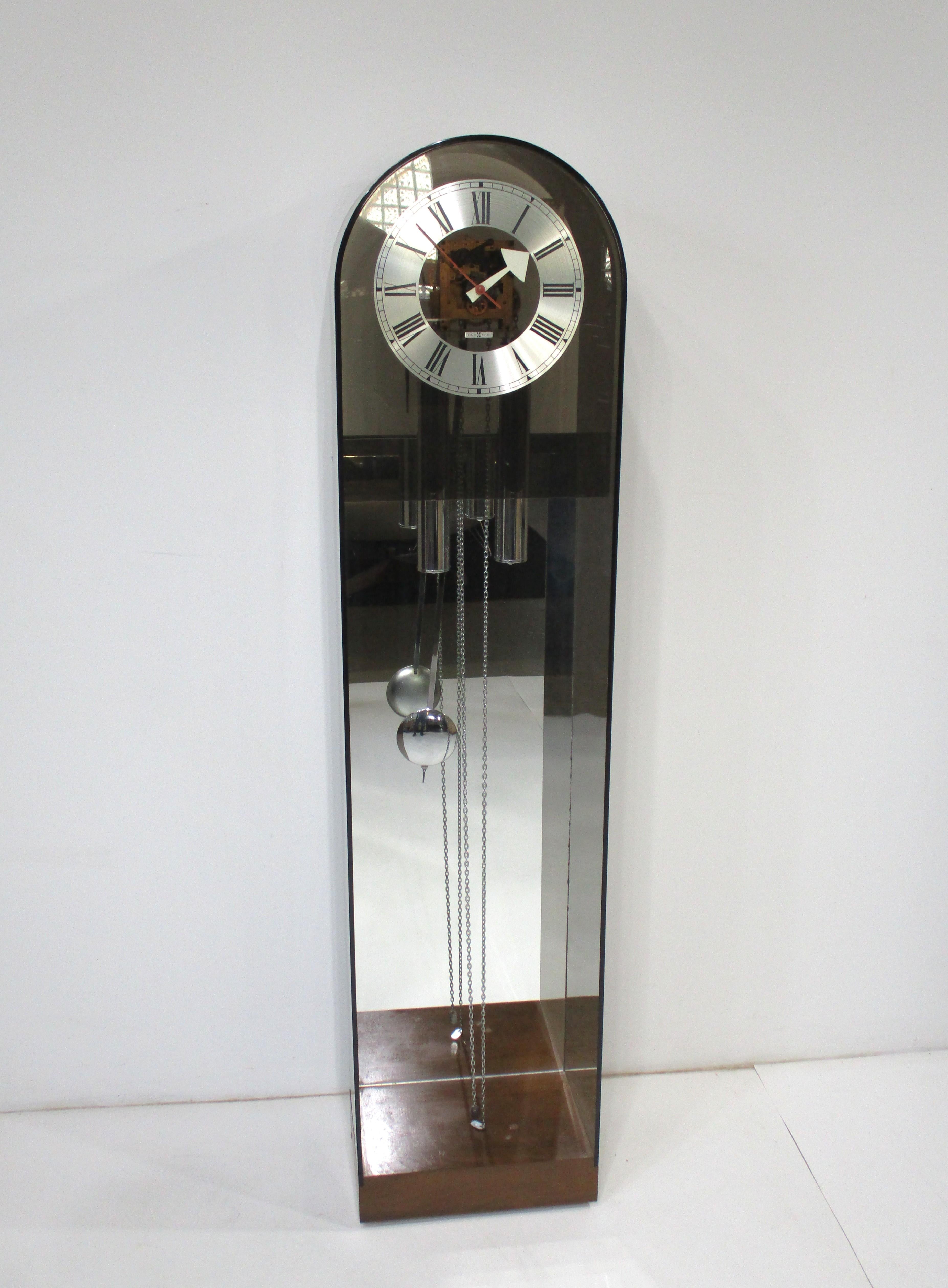 A newer sophisticated take on the grandfather clock with the case in smoked Lucite defining the outline of the clock. The face has a brushed aluminum face with mod hands in red and white marking the minutes and hours. The lower area has a cut out