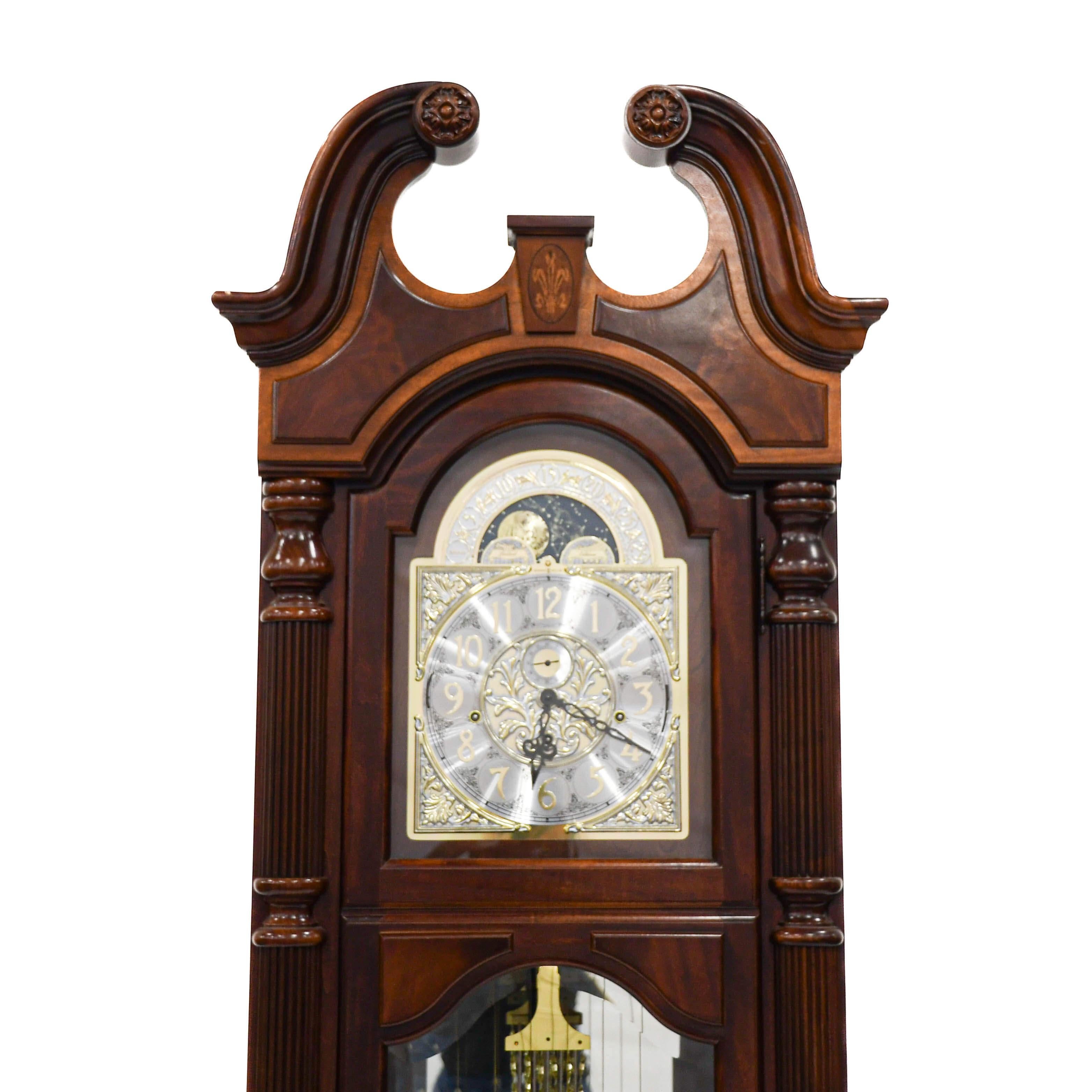 This Howard Miller Limited-Edition Grandfather Clock is an exquisite timepiece that showcases meticulous craftsmanship and ornate detail. The stately case, carved from rich wood, features a split pediment top with elegant scrollwork, while a