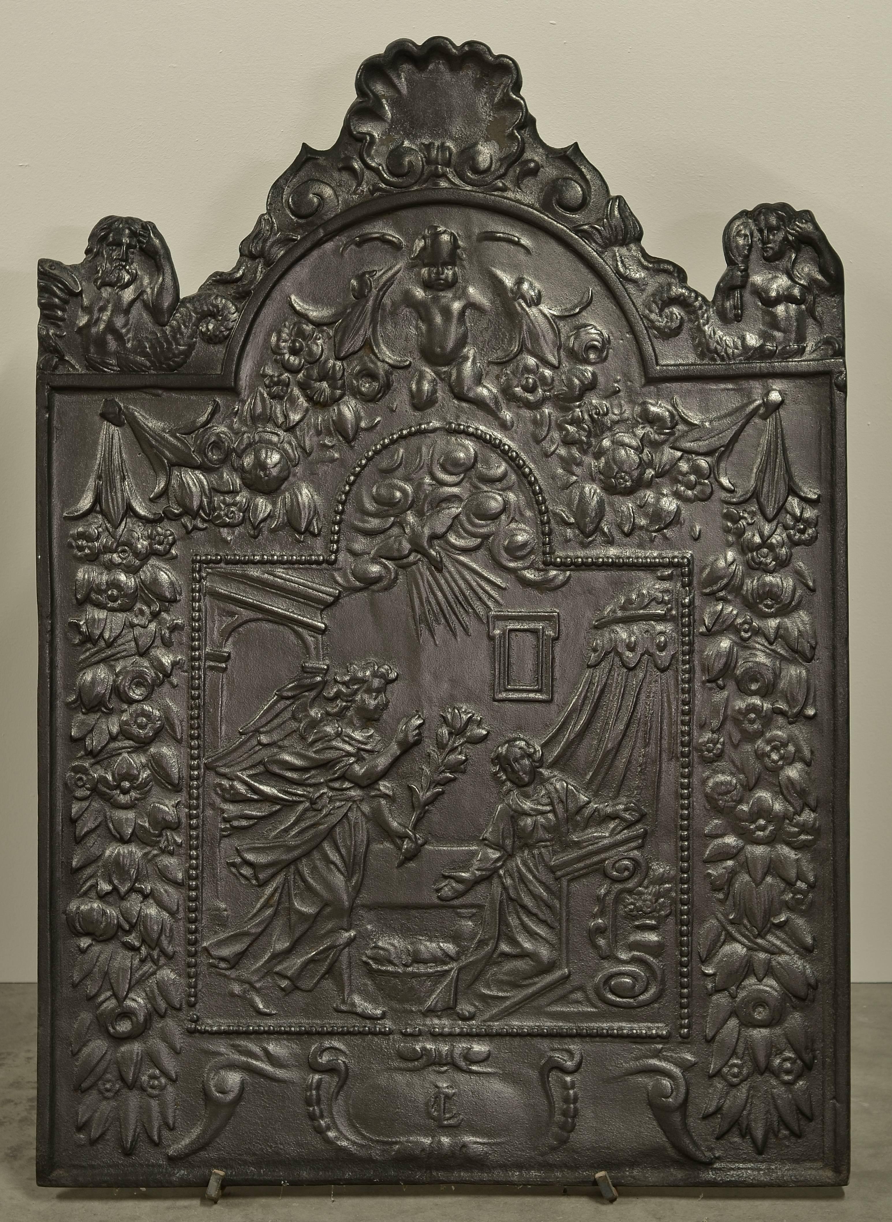 This huge one of a kind antique cast iron fireback displays the annunciation - the Christian celebration of the announcement by the angel Gabriel to the Virgin Mary that she would conceive and become the mother of Jesus, the Son of God, marking his