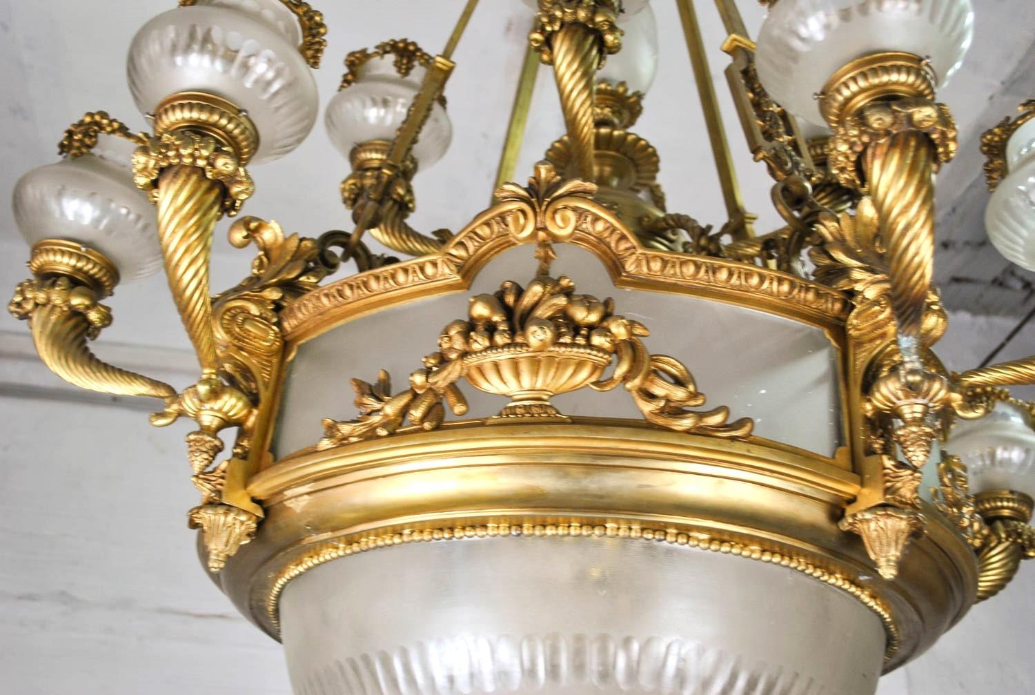Imponente lampadario in bronzo patinato e vetro smerigliato, composto da 13 luci, di cui 12 distribuite sui bracci. Ogni luce è contenuta in un tulipano di vetro smerigliato, ornato da grappoli d'uva pendenti. Anche la cintura e la coppa inferiore