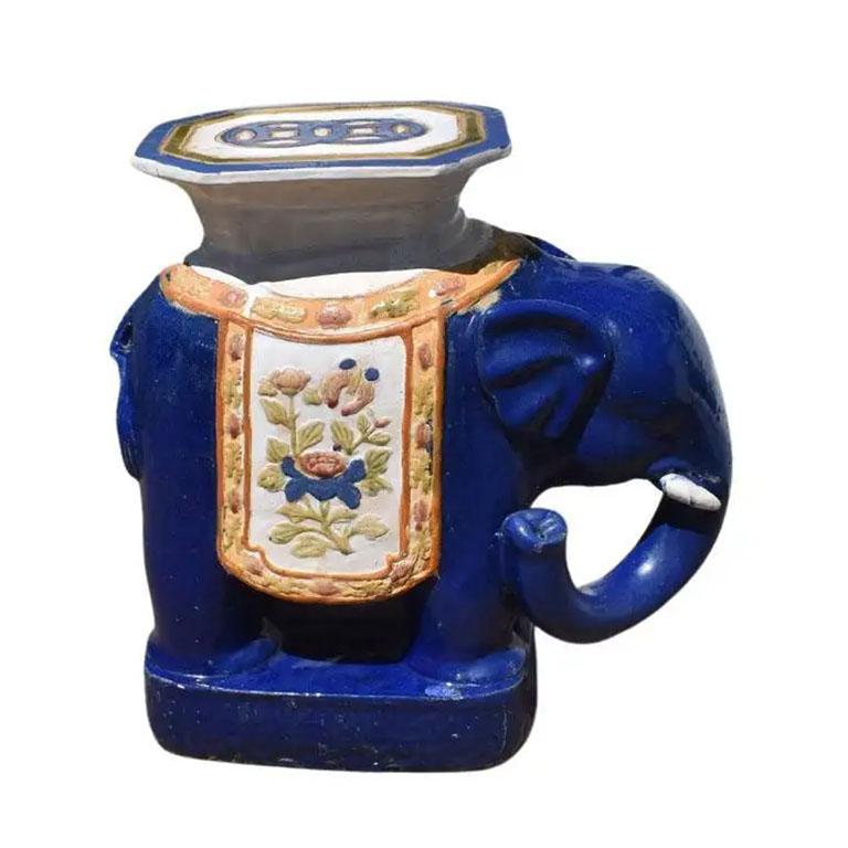 A rich blue, cream, green and pink ceramic elephant garden stool. This piece is tall and features an elephant shown with the trunk down and creamy white tusks. It wears a colorful pink, green and blue tapestry. At the top, is a chinoiserie design in