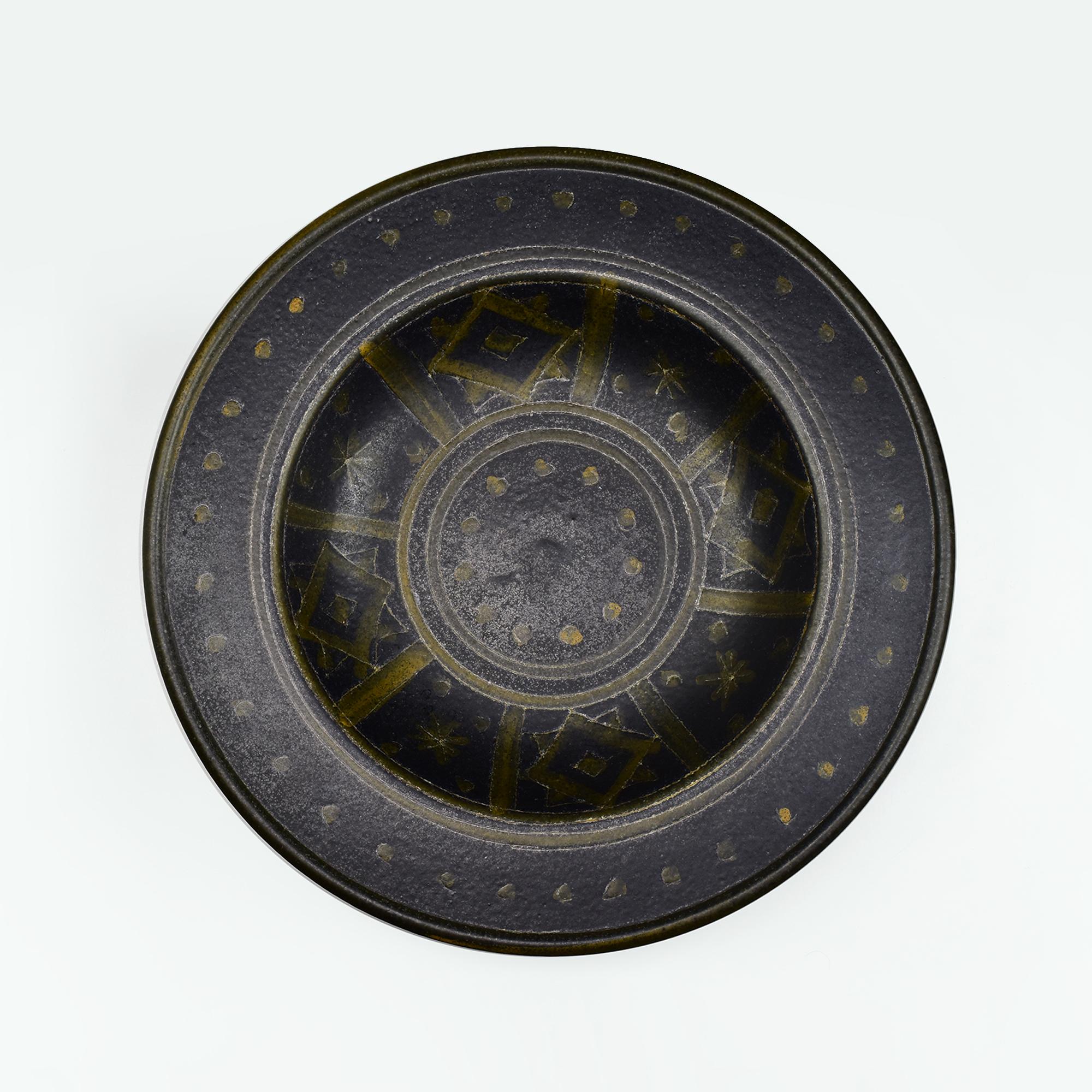 Questo sorprendente centrotavola XXL in ceramica presenta un audace disegno geometrico che ricorda gli iconici design di ICS e Gambone. La forma larga e poco profonda mostra un ricco smalto scuro ornato da intricati motivi geometrici gialli, tra cui
