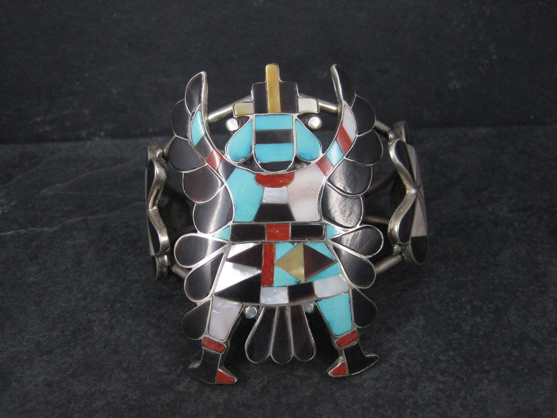 This gorgeous vintage knifewing bracelet is sterling silver.
It features inlay in onyx, white mother of pearl, yellow mother of pearl, coral and turquoise.

It is the creation of master Zuni silversmith Annalee Tekala.

The face of this bracelet