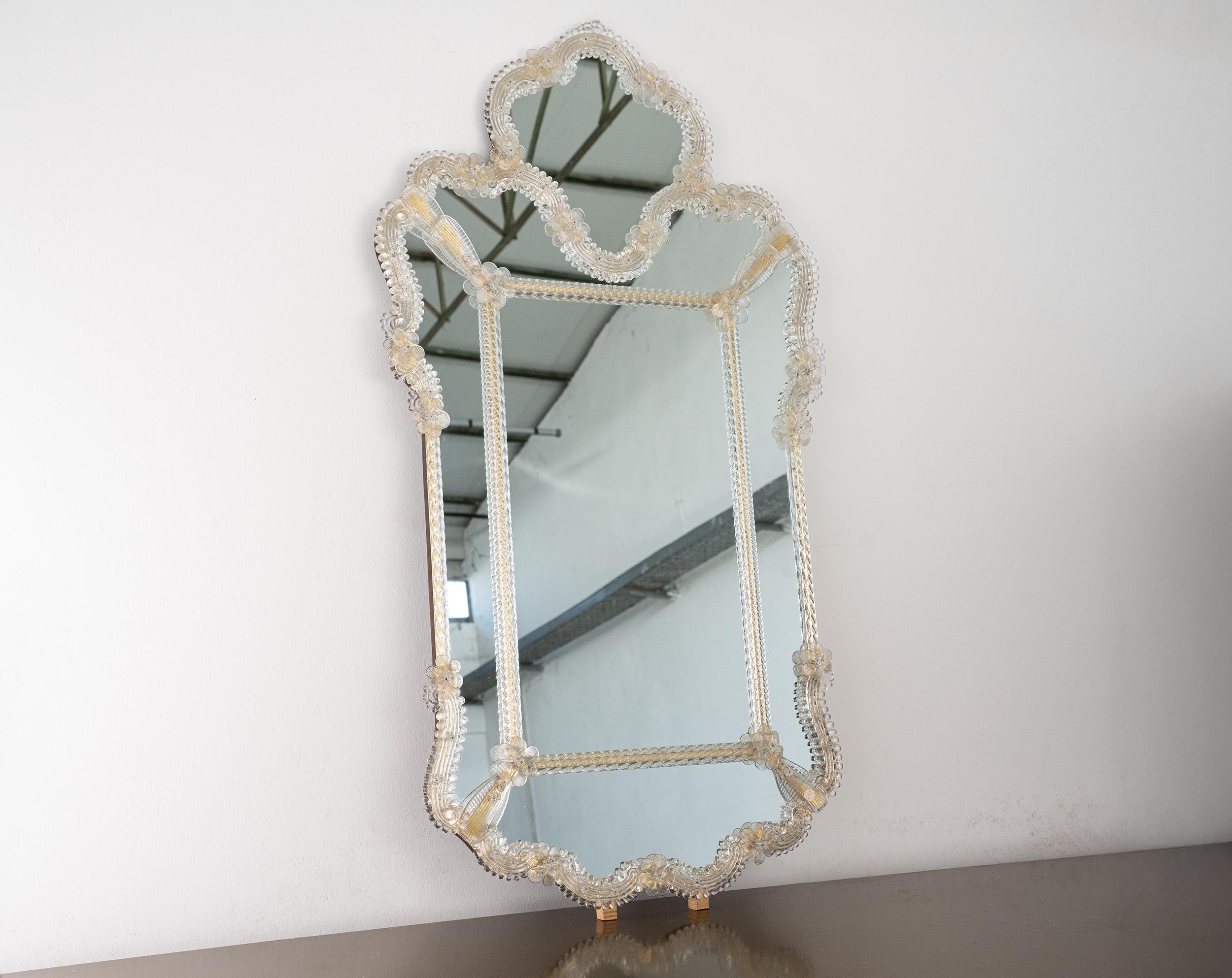 Magnifique miroir mural vénitien vintage entièrement réalisé à la main en verre de Murano, présentant des rubans de verre clair et de délicats détails floraux infusés d'or.

Le miroir est doté d'un support en bois avec une boucle de suspension, prêt