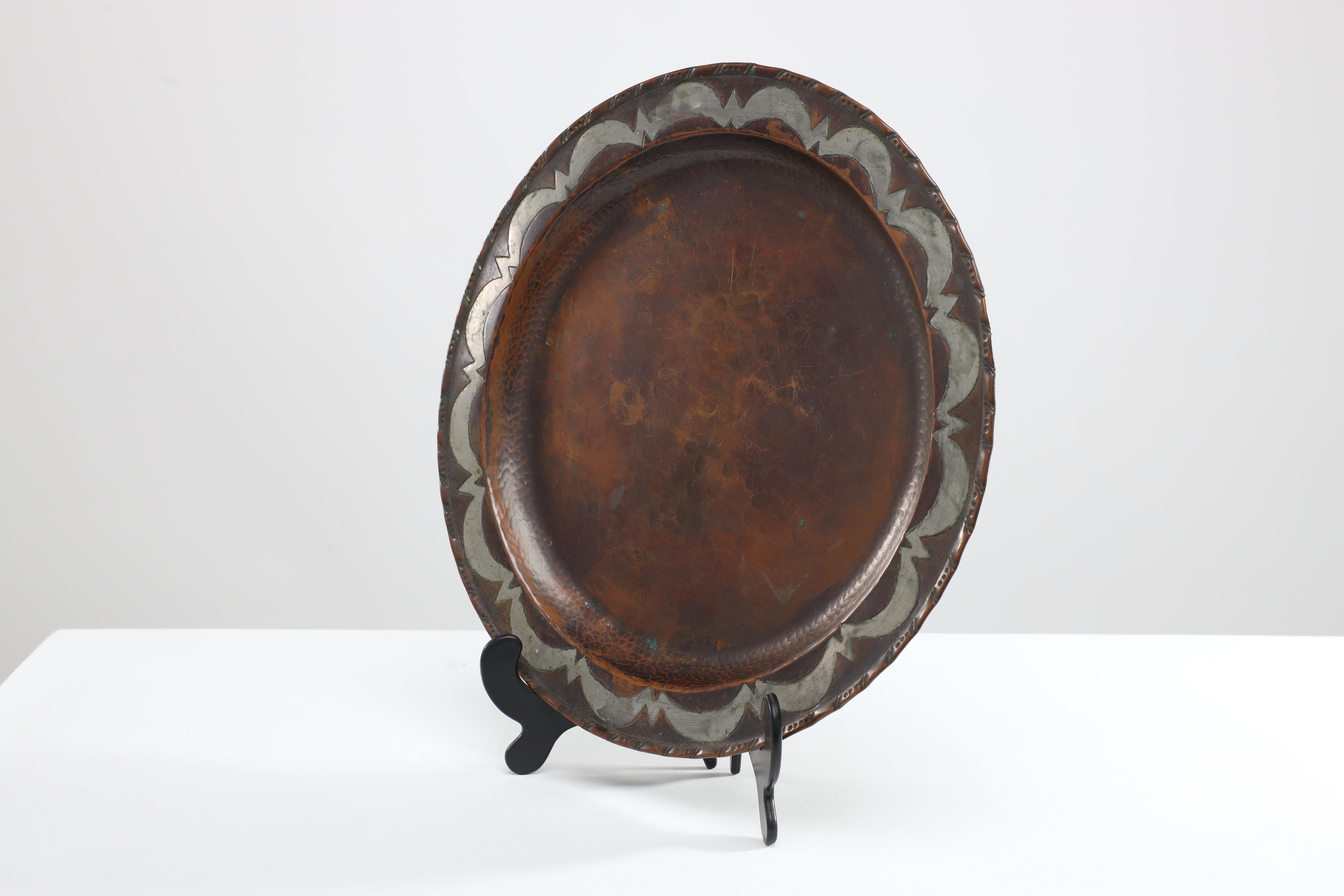 Hugh Wallis. An Arts & Crafts hand formed round copper plate with chased decoration to the edge and inlaid to the inner edge with pewter. Stamped HW in a square. Nice chocolait patina. Hugh Wallis was based in Altrincham, near Manchester in the