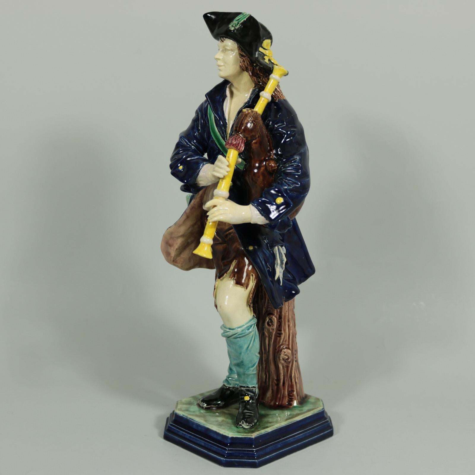 Hugo Lonitz Majolica figure which features a young man playing bagpipes, bottle in pocket. Colouration: blue, brown, yellow, are predominant. The piece bears maker's marks for the Lonitz pottery. Bears a pattern number, '1337'.