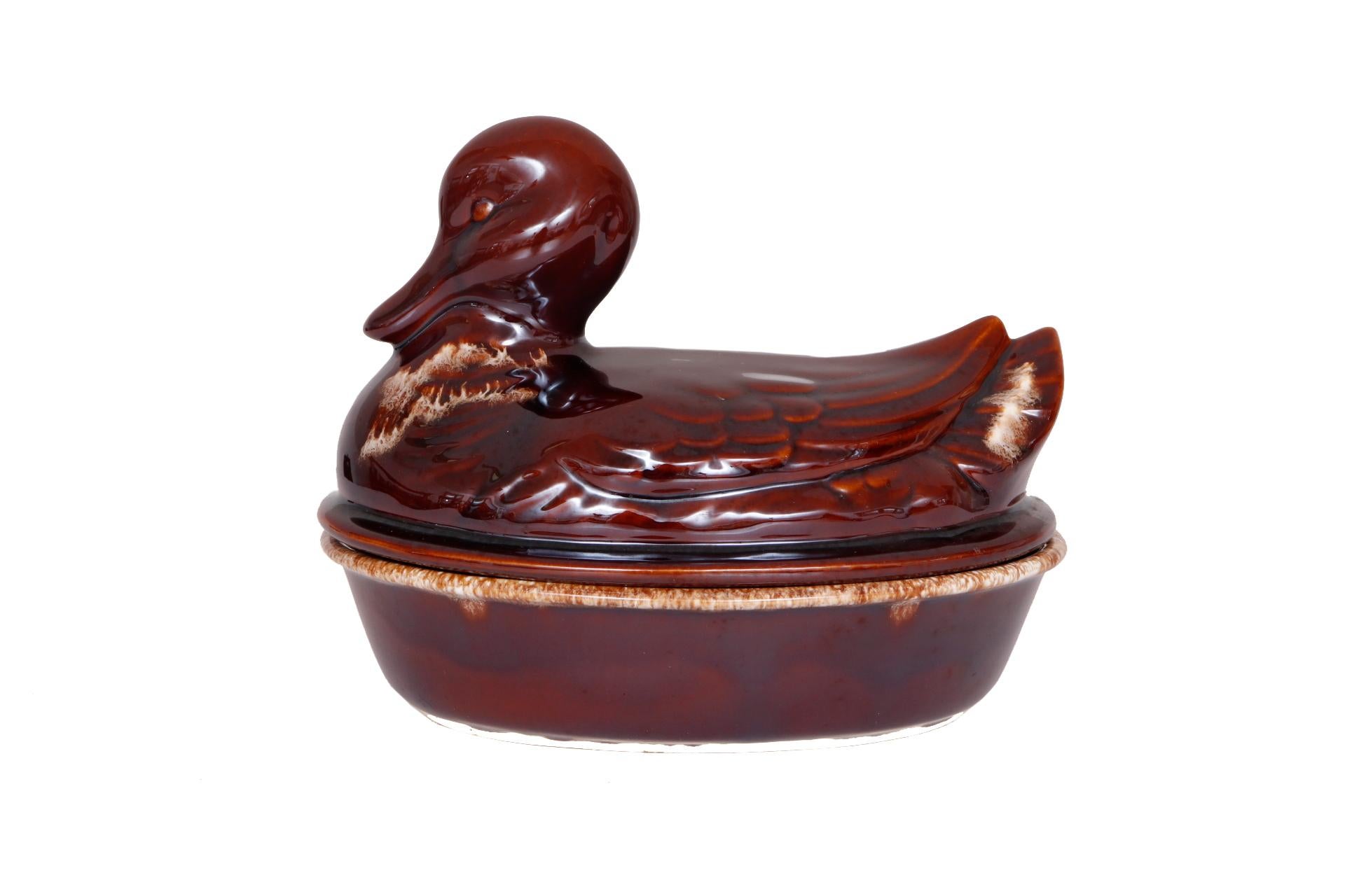 A duck shaped serving dish made by Hull Pottery and Co. An oval dish and duck shaped lid are decorated in a rich brown drip glaze. The duck is pressed with eye and feather details. Marked underneath “H.P. Co Oven Proof USA”.