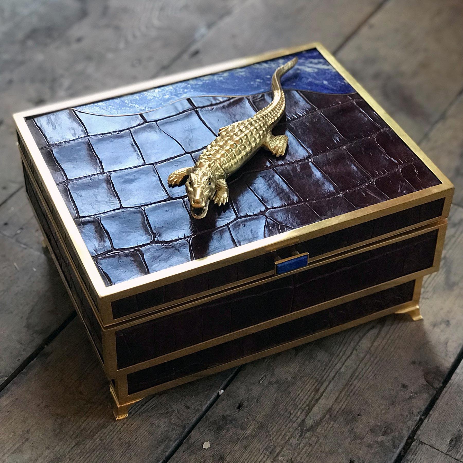 Ein beeindruckender Feuchtmittel von Glynn Lockett, der mit Krokodillederhaut und vergoldeten Beschlägen versehen ist.

Die Oberseite ist mit einem Krokodil mit Saphiraugen verziert, neben einer wellenförmigen Lapislazuliplatte, die einen Fluss