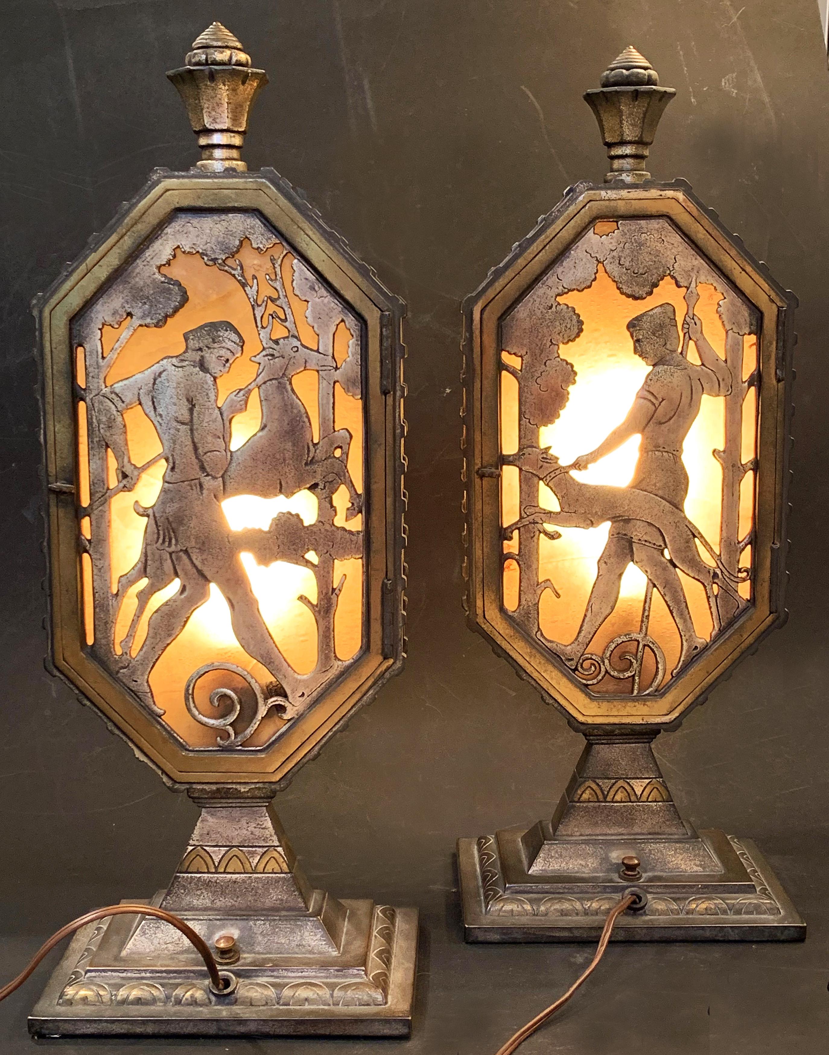 Rare and gorgeously crafted, this pair of illuminated bronze lamps depict a pair of hunters in Medieval garb, walking their hounds through the deep woods, as if in an old fairy tale or legend. The pierced bronze scenes, in silvered and patinated