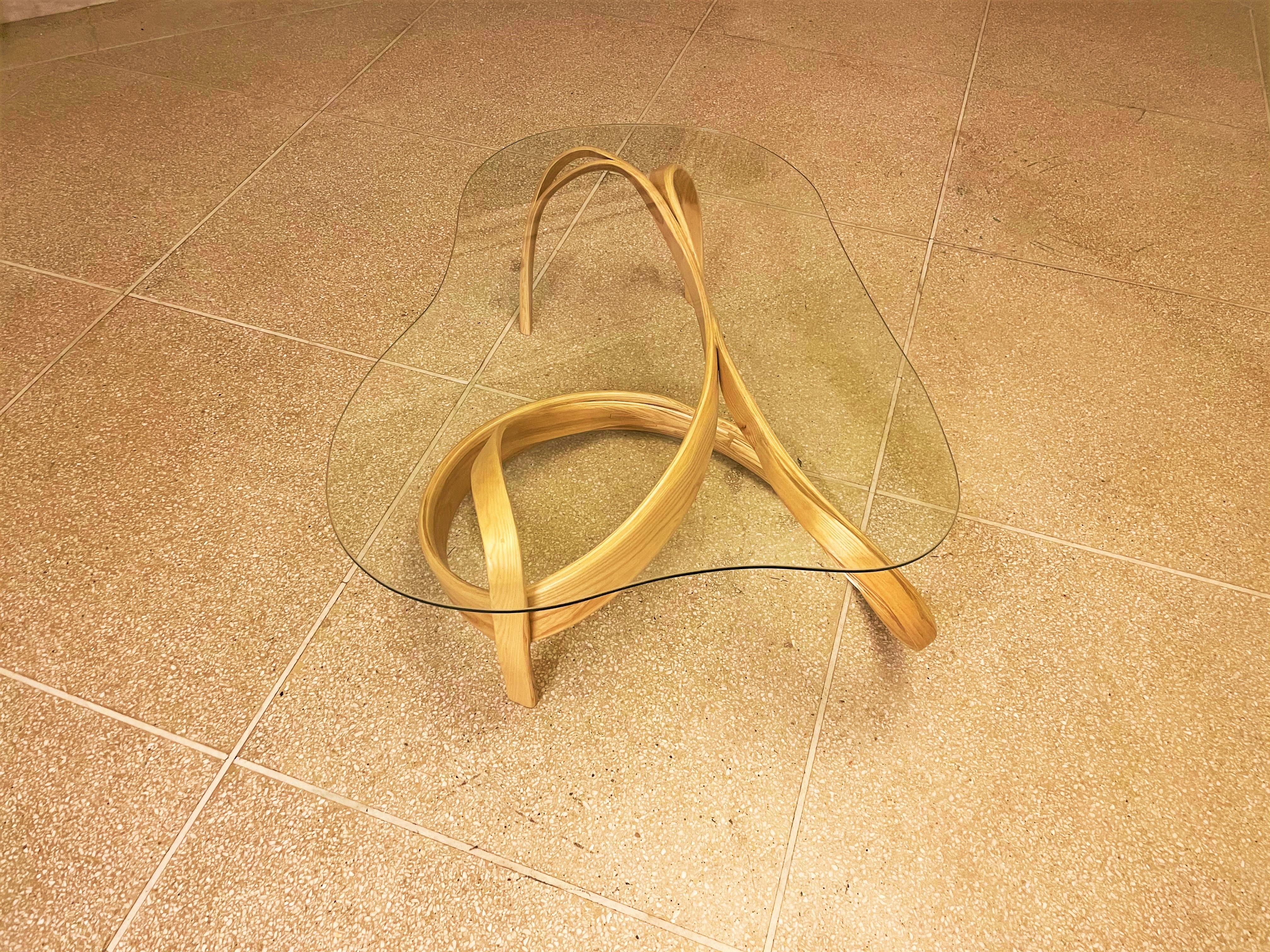 La table de cocktail 101 est fabriquée en pliant du bois de frêne pour créer le cadre de base qui équilibre un plateau en verre transparent qui est incurvé pour compléter les courbes du cadre. Le bois qui coule en continu s'étend en largeur, ce qui