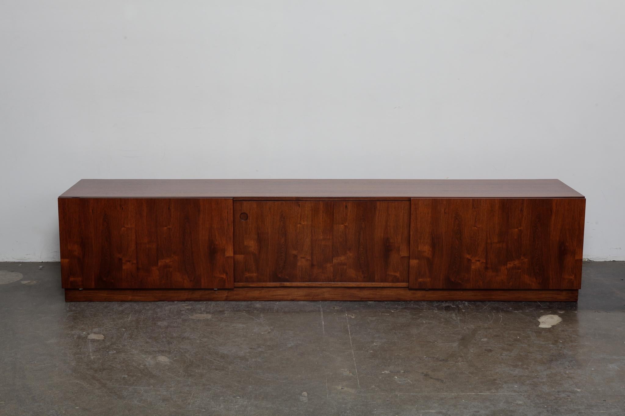 Long, very low rosewood sideboard, Danish, by Ib Kofod Larsen, produced by Faarup Mobelfabrik. It has 3 sliding doors with center drawers, sitting on a plinth base. Excellent grain throughout, newly refinished in lacquer.