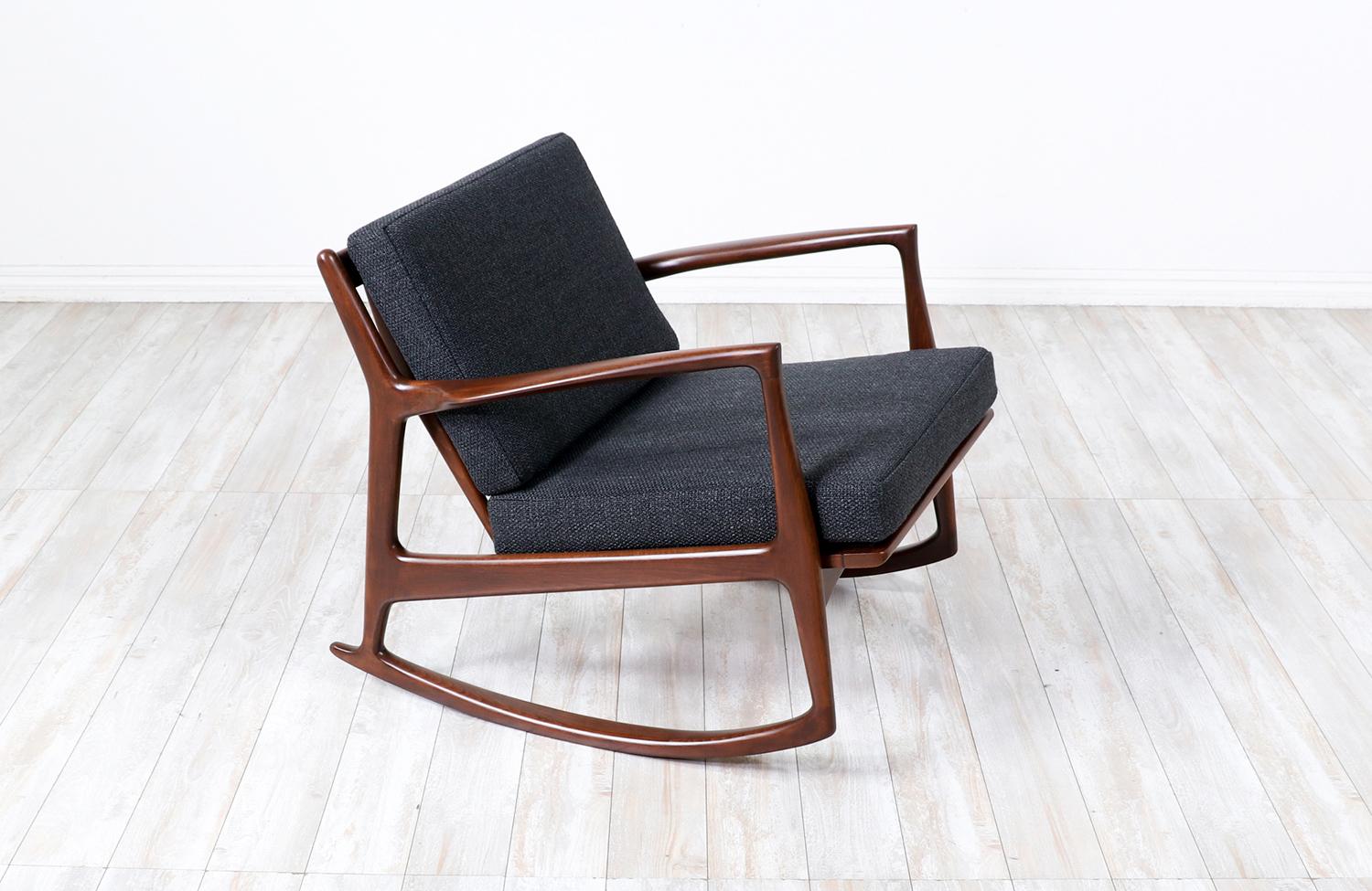Stunning rocking chair designed by Danish furniture designer Ib-Kofod Larsen for Selig in Denmark c. 1960’s. Featuring a walnut-stained beechwood frame with a slatted back. New high-density foam cushions are newly reupholstered in a dark blue tweed