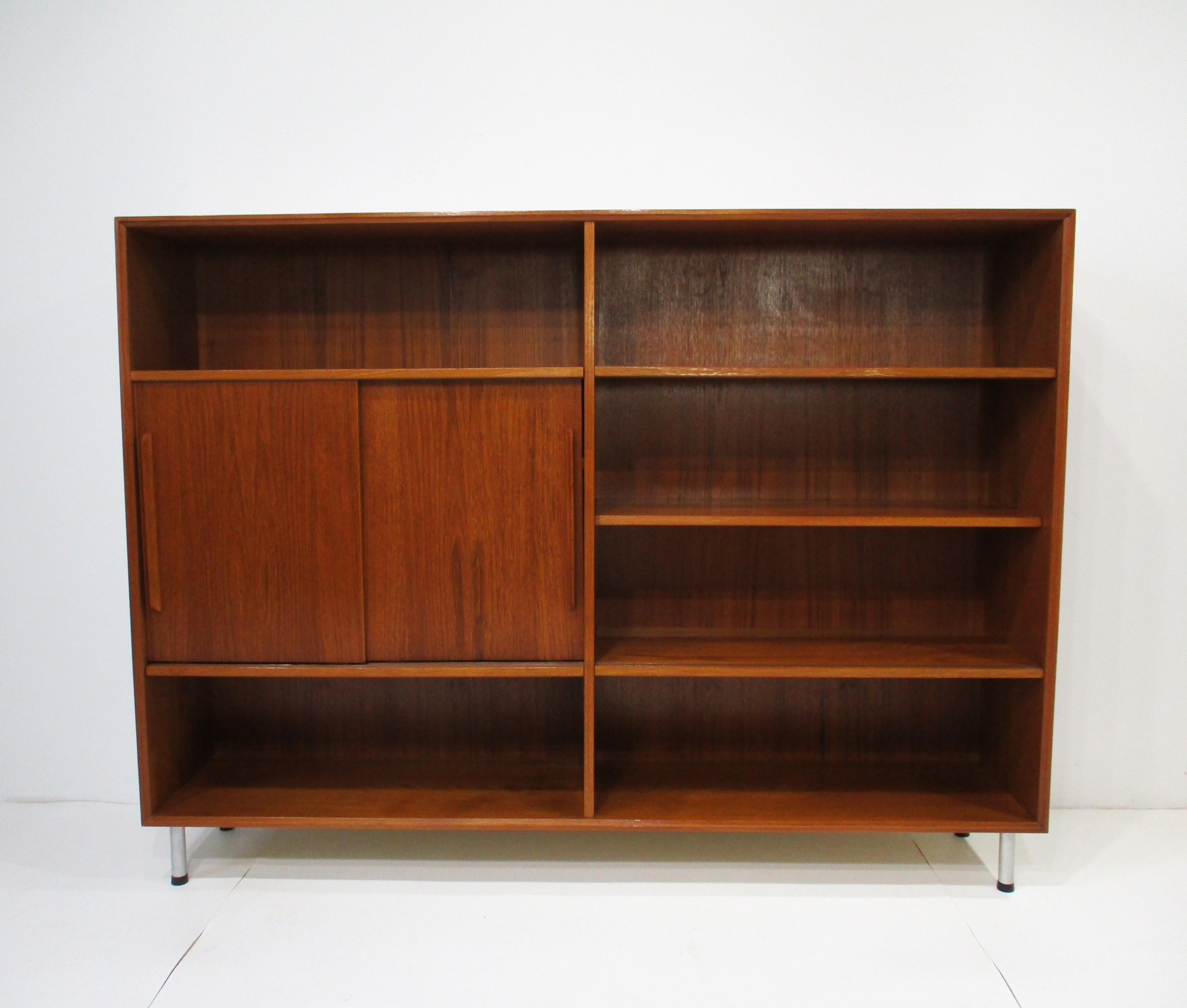 Una libreria in legno di teak di ottima fattura con doppie ante scorrevoli, due ante scorrevoli in vetro e due ripiani regolabili. Poggiato su gambe in alluminio con pad per proteggere il pavimento, è un pezzo robusto e molto funzionale. Prodotto in