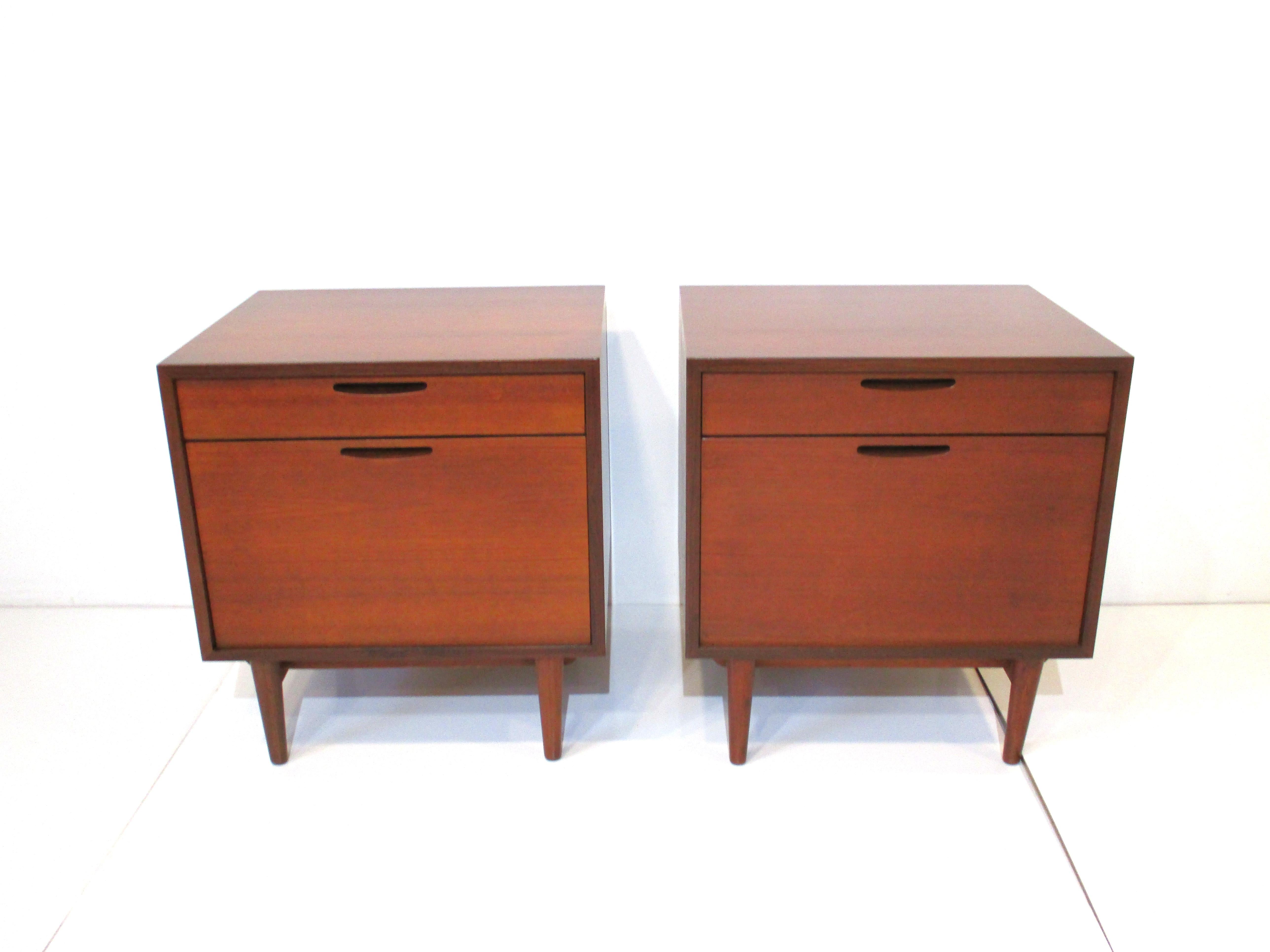 A matching pair of rare IB Kofod - Larsen teak wood nightstands / side table cabinets with top drawer and flap down lower door with storage. The flap door has brass hinges and contrasting wood grain giving the piece a wonderful design aesthetic, a