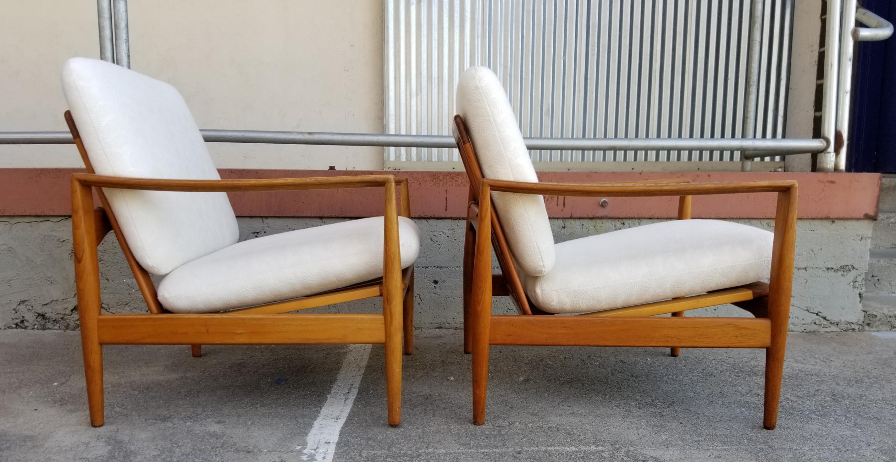 A pair of teak lounge chairs designed by Ib Kofod-Larsen and sold through notable retailer Illum Bologhus, Copenhagen. Crafted in solid teak with unusual finger-joint detail at front of arms. Retaining Illum Bologhus metal label on each chair.
